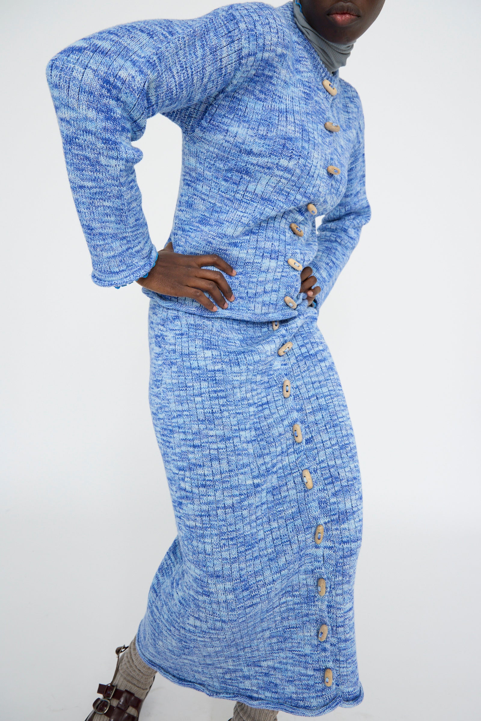 Person in a Twisted Cotton Drip Cardigan by Baserange in Blue Melange, paired with a matching skirt adorned with wooden buttons, poses against a plain background with hands on their hips. 