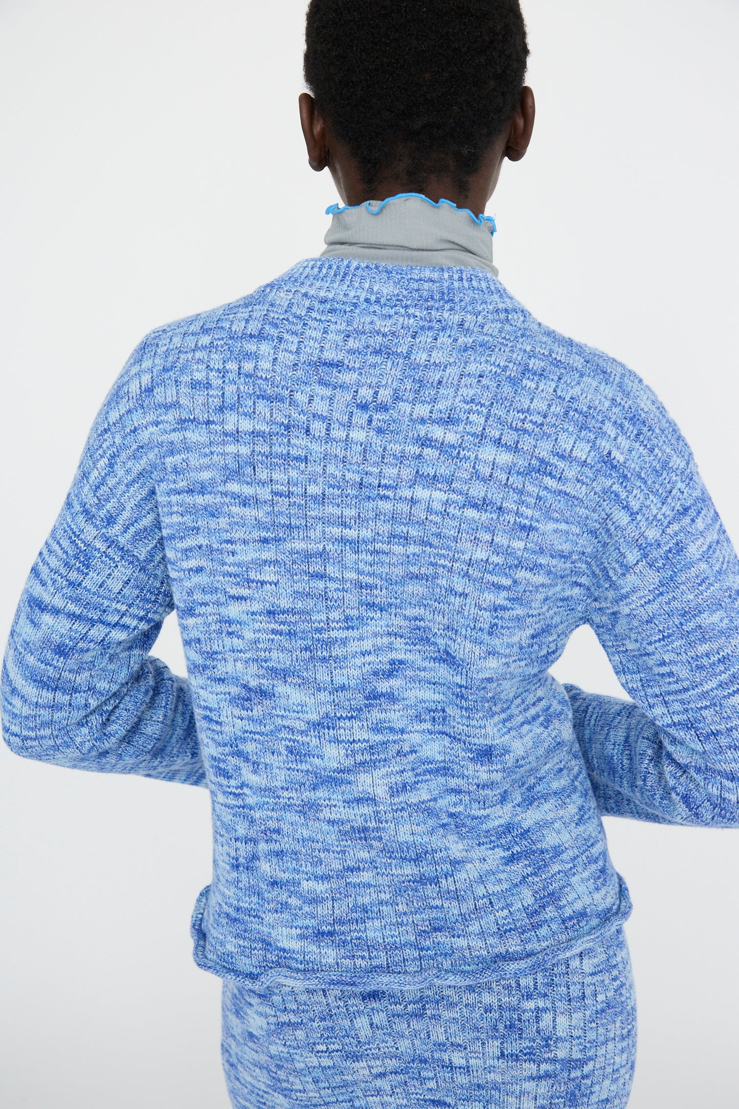 A model in a blue melange Twisted Cotton Drip Cardigan by Baserange and a matching skirt stands with their back to the camera against a plain background.