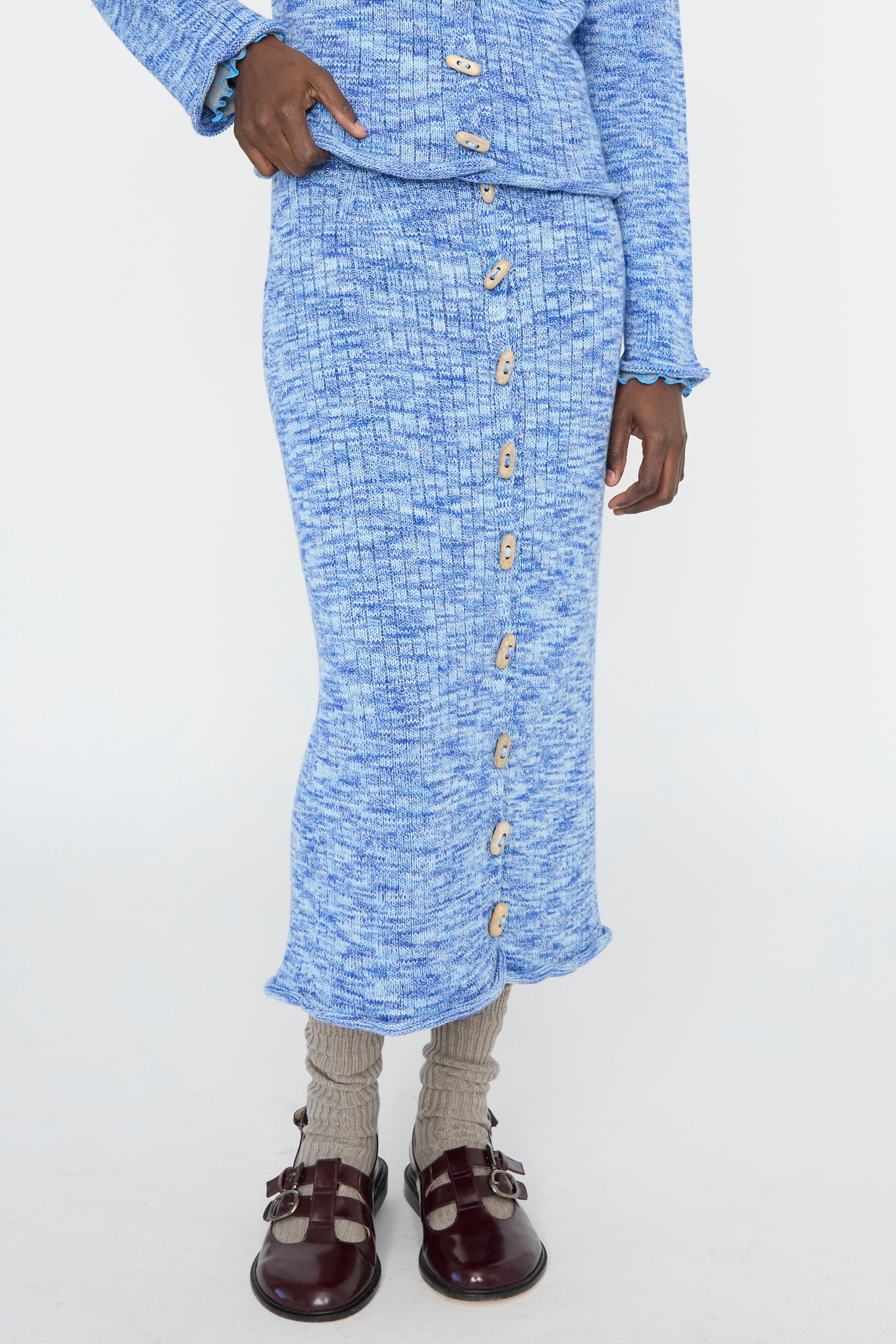 Dressed in Baserange's blue melange Twisted Cotton Drip Skirt, paired with a ribbed knit top featuring large buttons against a white backdrop. 