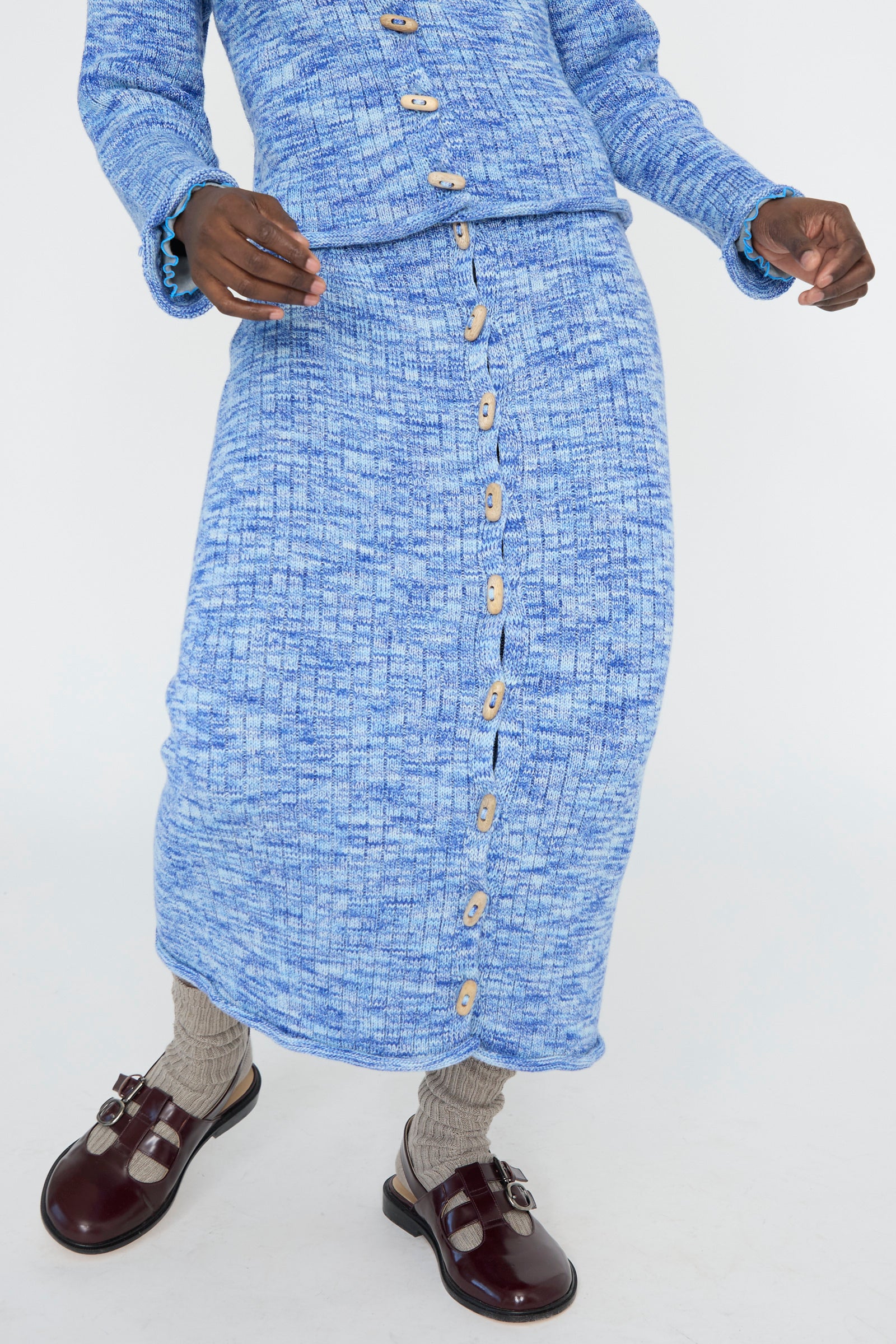 A person wearing a blue melange knit button-up top and Baserange's Twisted Cotton Drip Skirt in Blue Melange, complemented by gray socks and brown shoes against a white backdrop. 