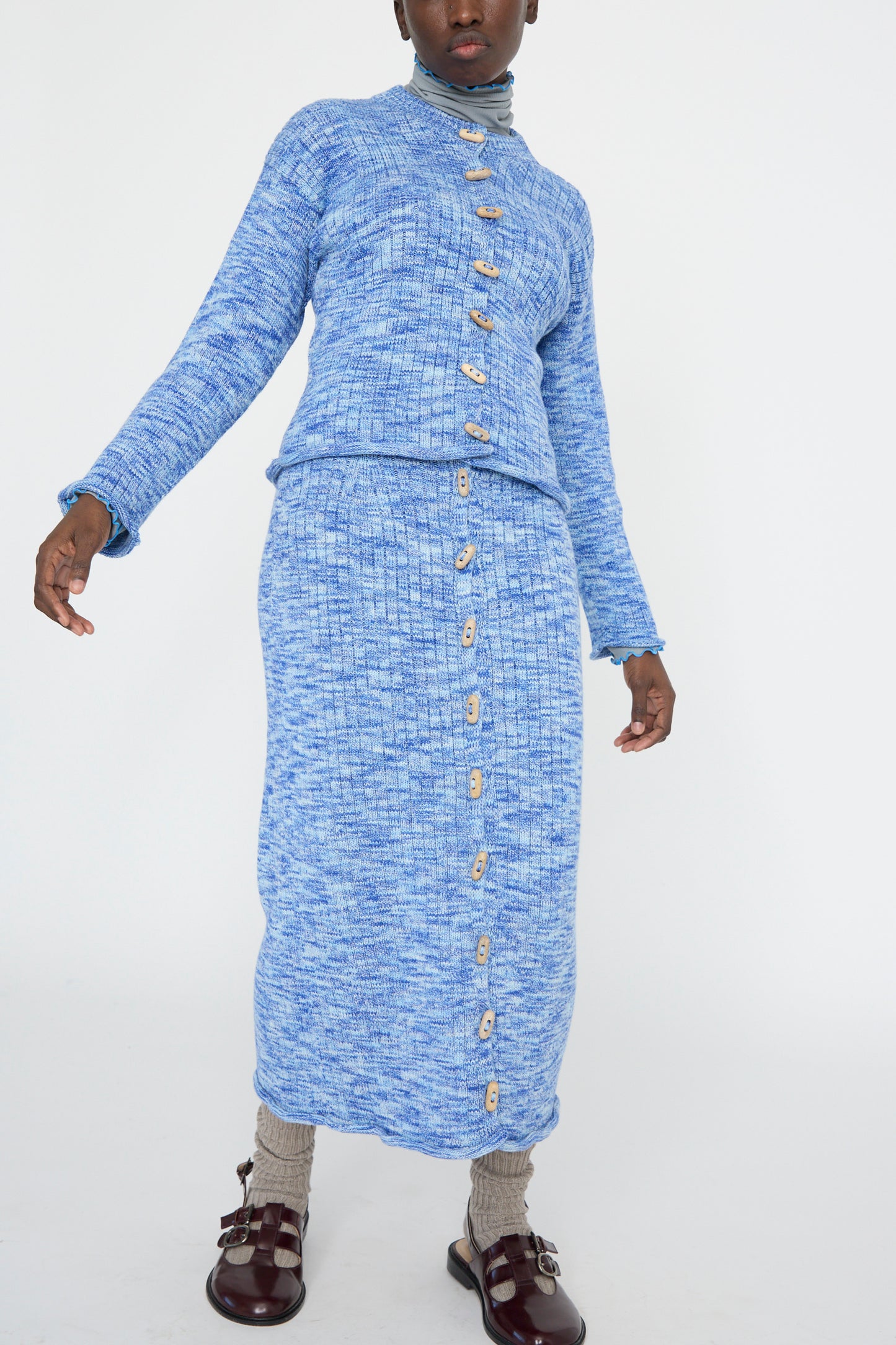 A person is wearing the Twisted Cotton Drip Skirt in Blue Melange by Baserange, a blue melange knit button-up sweater set crafted from organic cotton, paired with brown shoes and gray socks, standing against a white background.