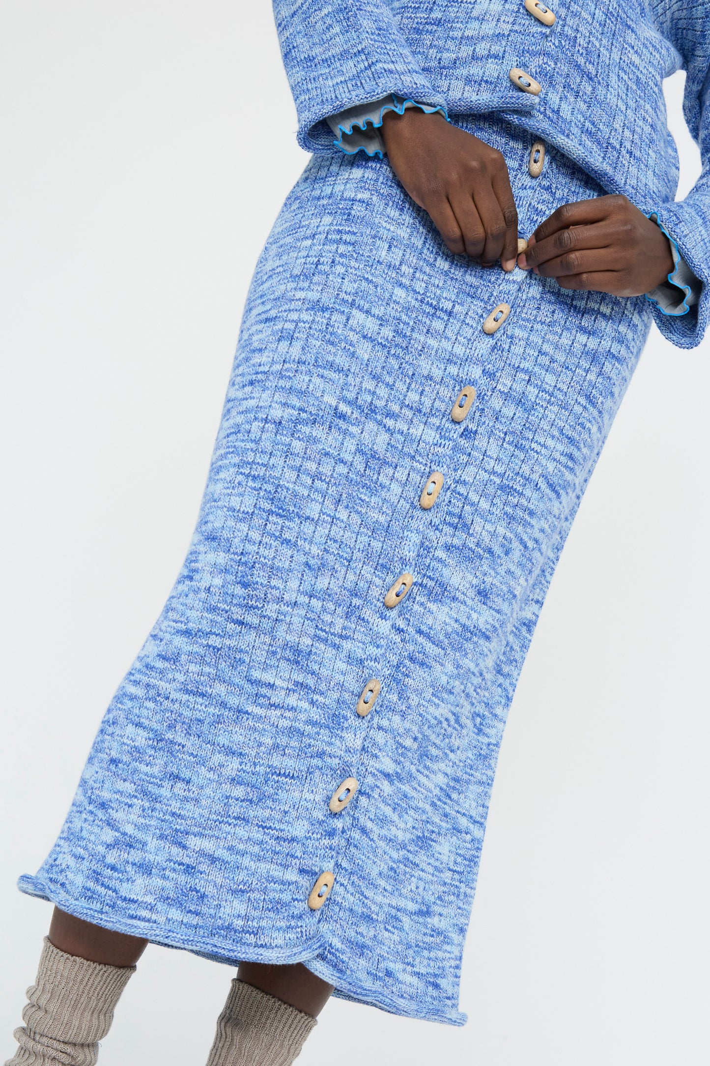 The Baserange Twisted Cotton Drip Skirt in Blue Melange, featuring wooden buttons and an elastic waist, is paired with a matching top and cozy gray socks for effortless style and sustainability.