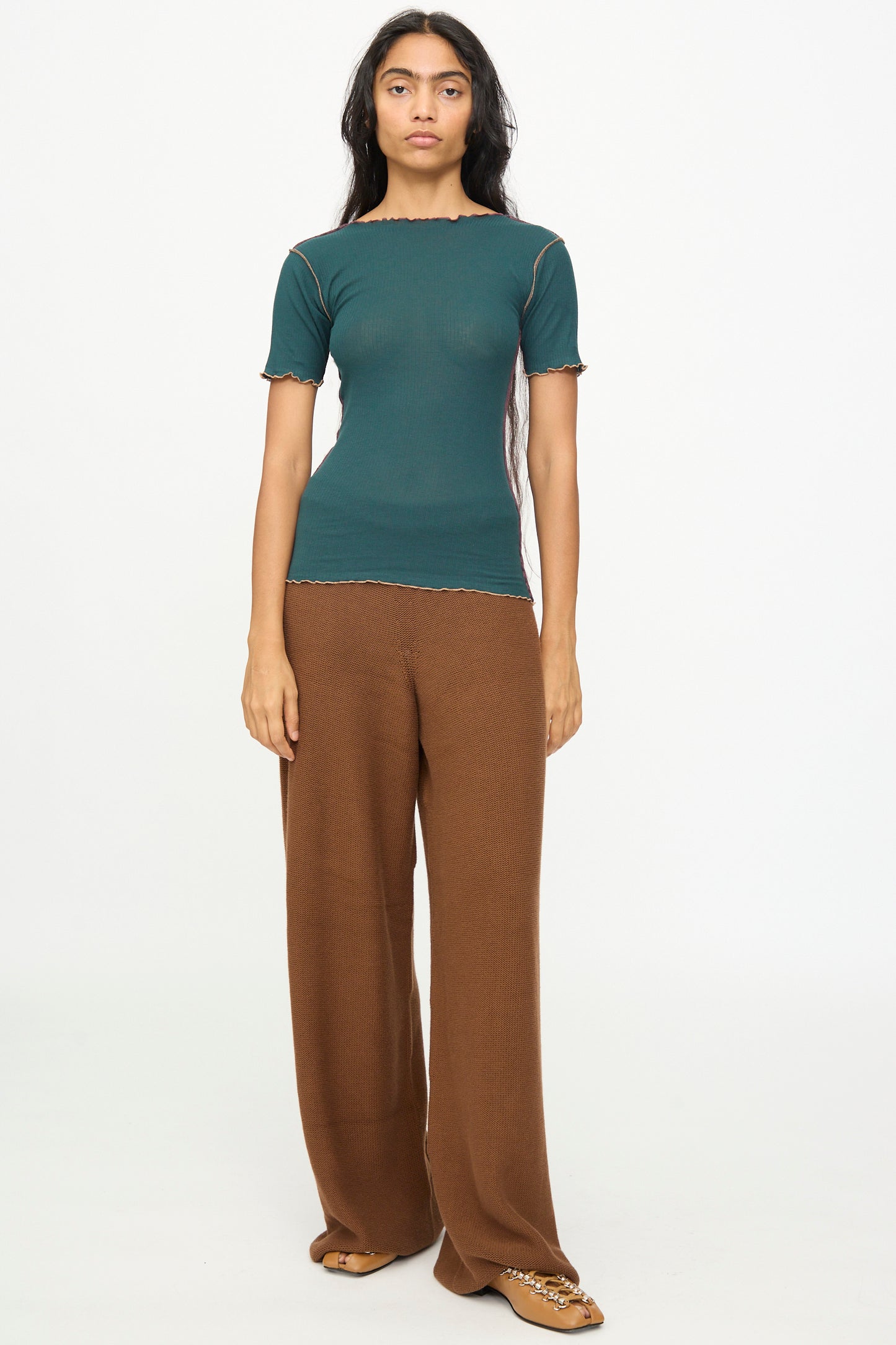 A woman with long dark hair and a neutral expression stands against a plain background, wearing the Baserange Vein Tee Shirt in Hirsu Green—a soft cotton rib, short-sleeve boat neck top—styled with loose brown pants and brown shoes.