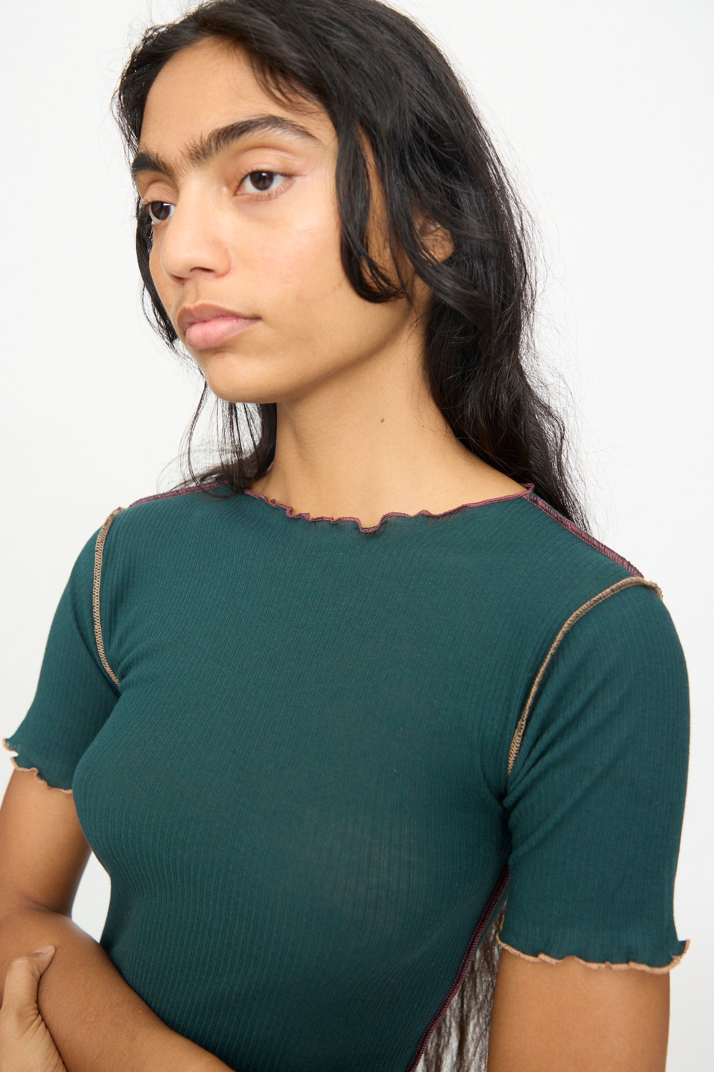A person with long dark hair wears the Baserange Vein Tee Shirt in Hirsu Green—a fitted, short-sleeve cotton rib top with lettuce edge seams—standing against a plain light background.