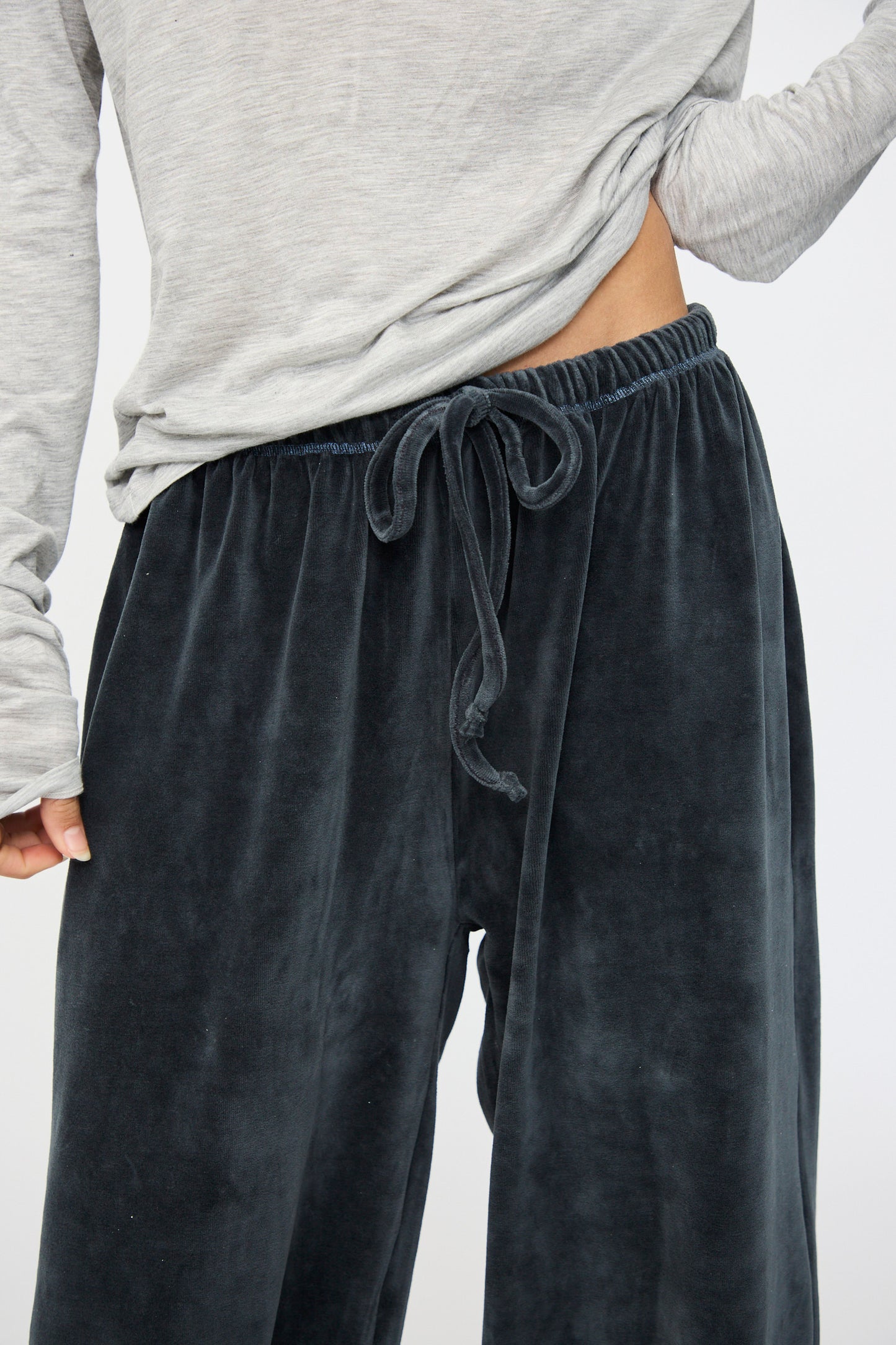A person wearing the Baserange Velour Pillow Pant in Petroleum Blue, with a light gray long sleeve top slightly lifted to reveal the pant’s waistband.