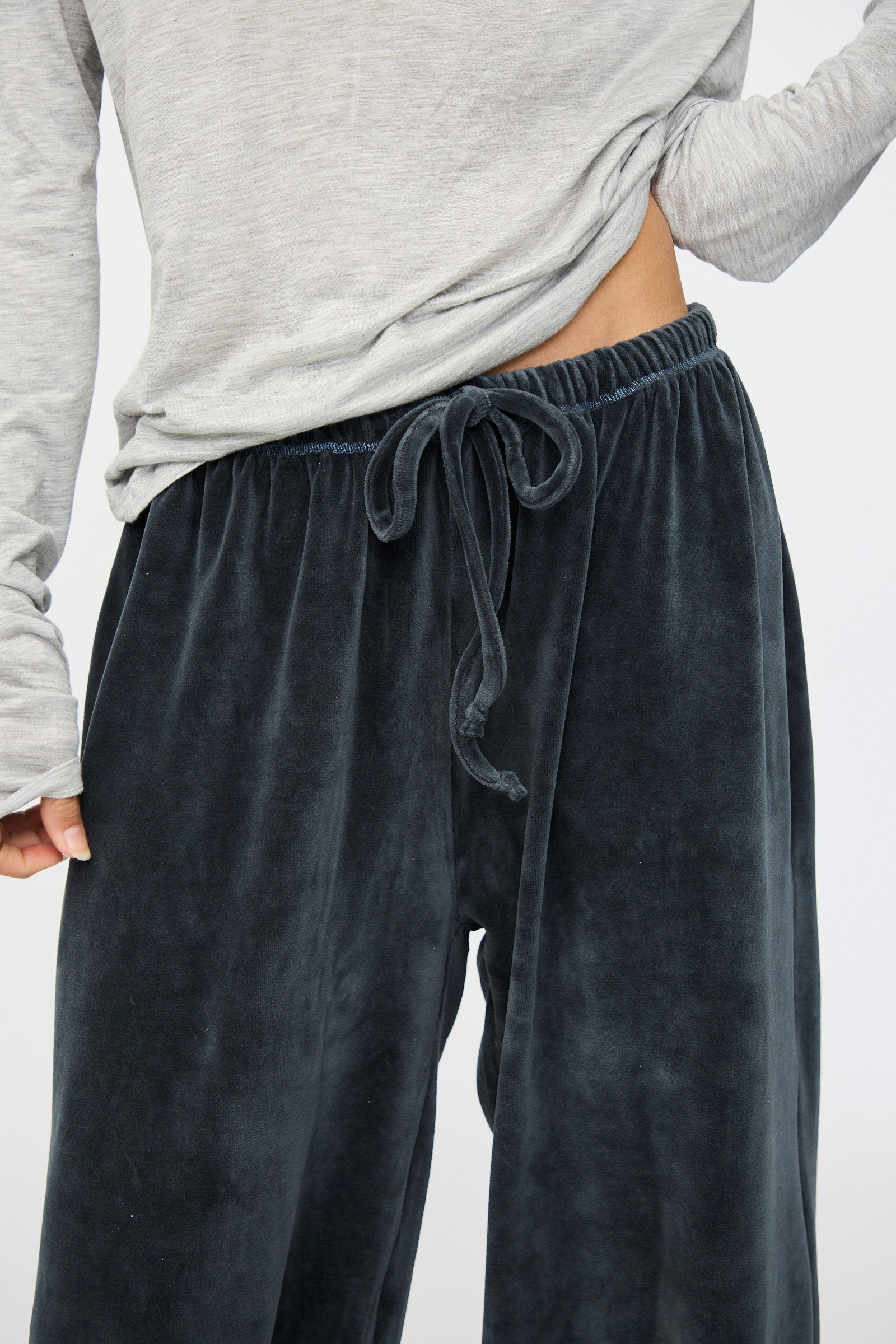 A person wearing the Baserange Velour Pillow Pant in Petroleum Blue, with a light gray long sleeve top slightly lifted to reveal the pant’s waistband.