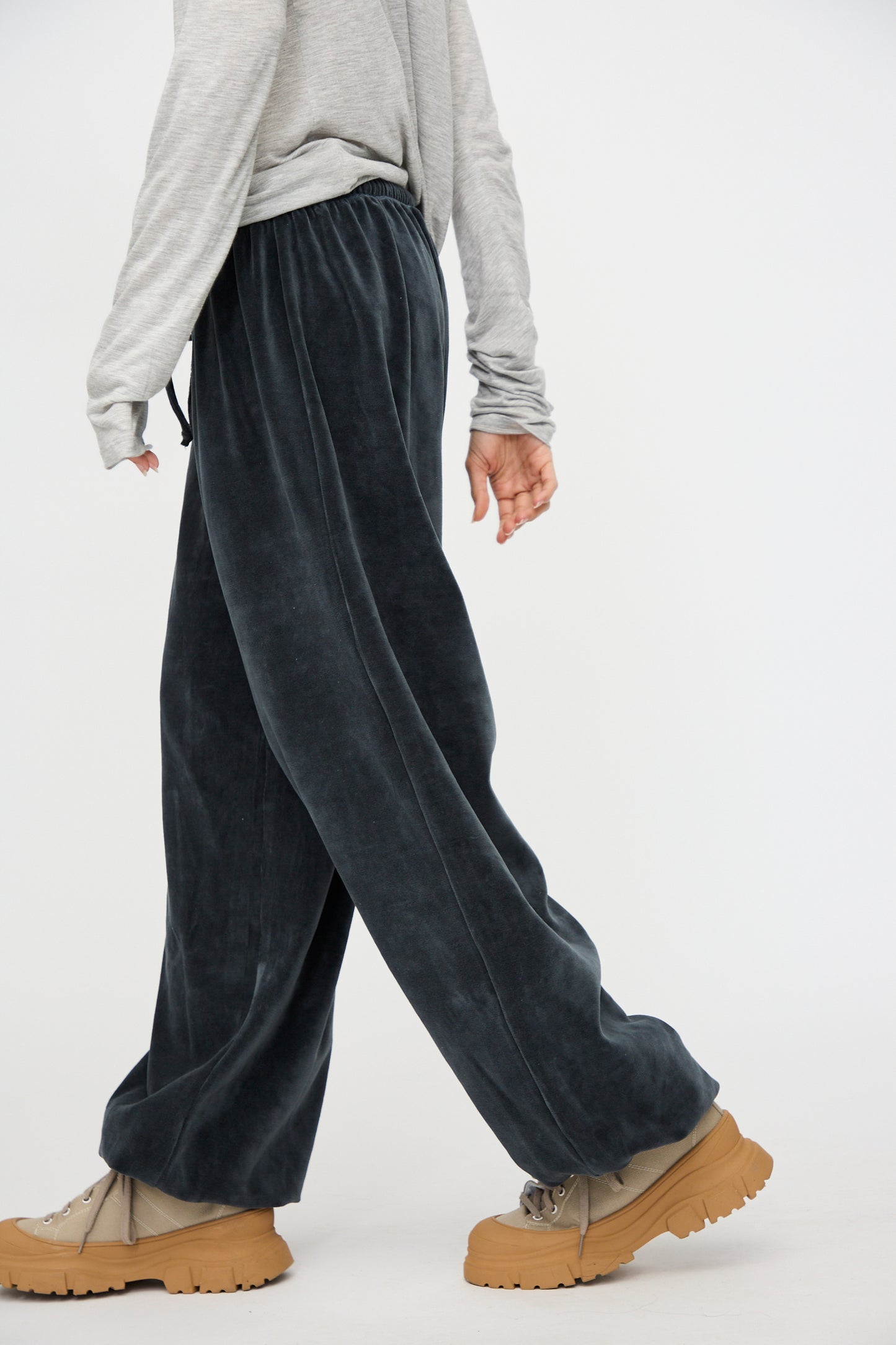 A person wearing a light gray recycled cotton long-sleeve shirt, Baserange Velour Pillow Pant in Petrolium Blue, and chunky tan shoes, walks with one arm at their side against a plain background.