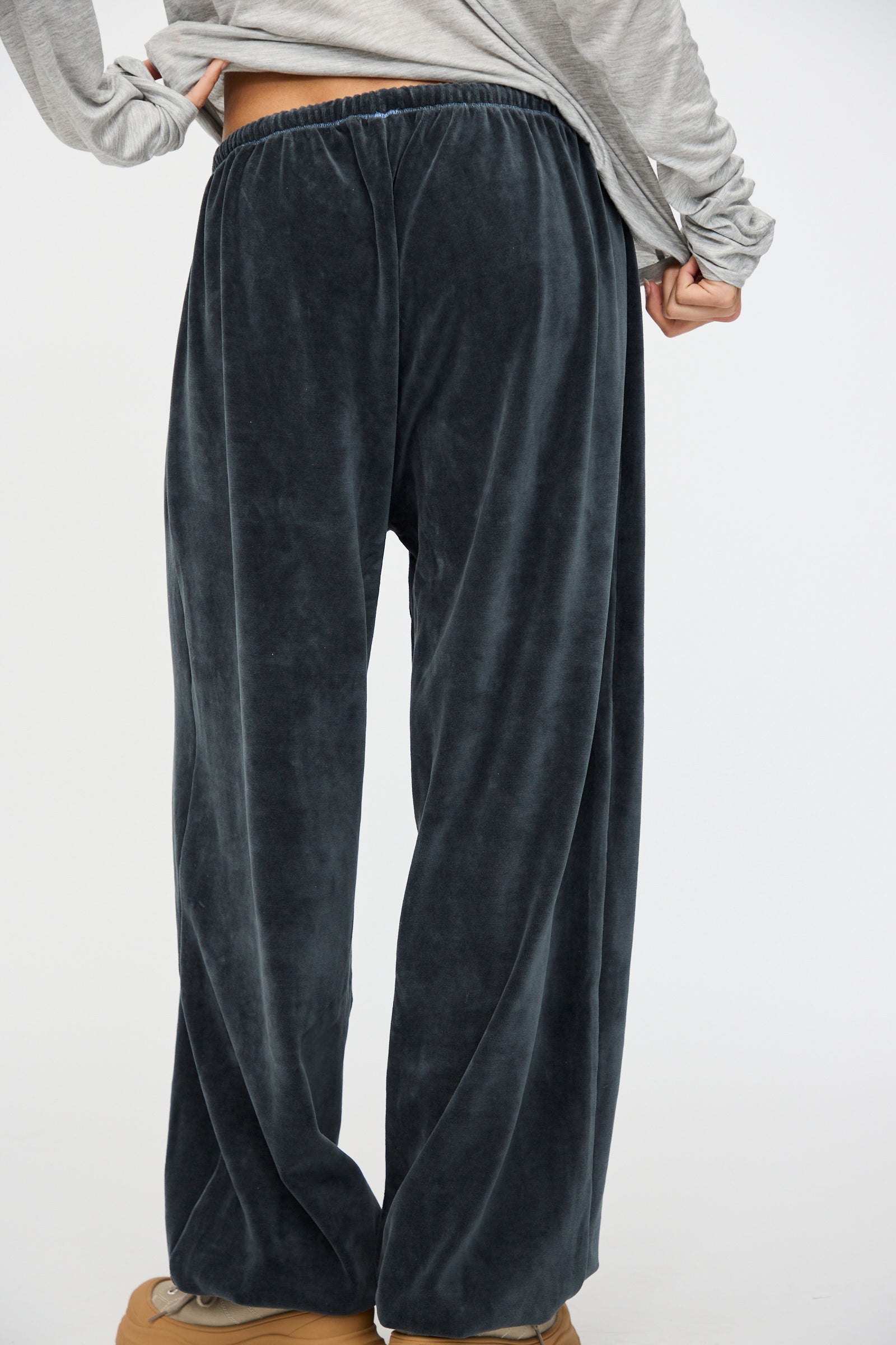 A person wearing Baserange's Velour Pillow Pant in Petrolium Blue, a light gray long-sleeve shirt, and tan shoes stands with one hand on their hip.
