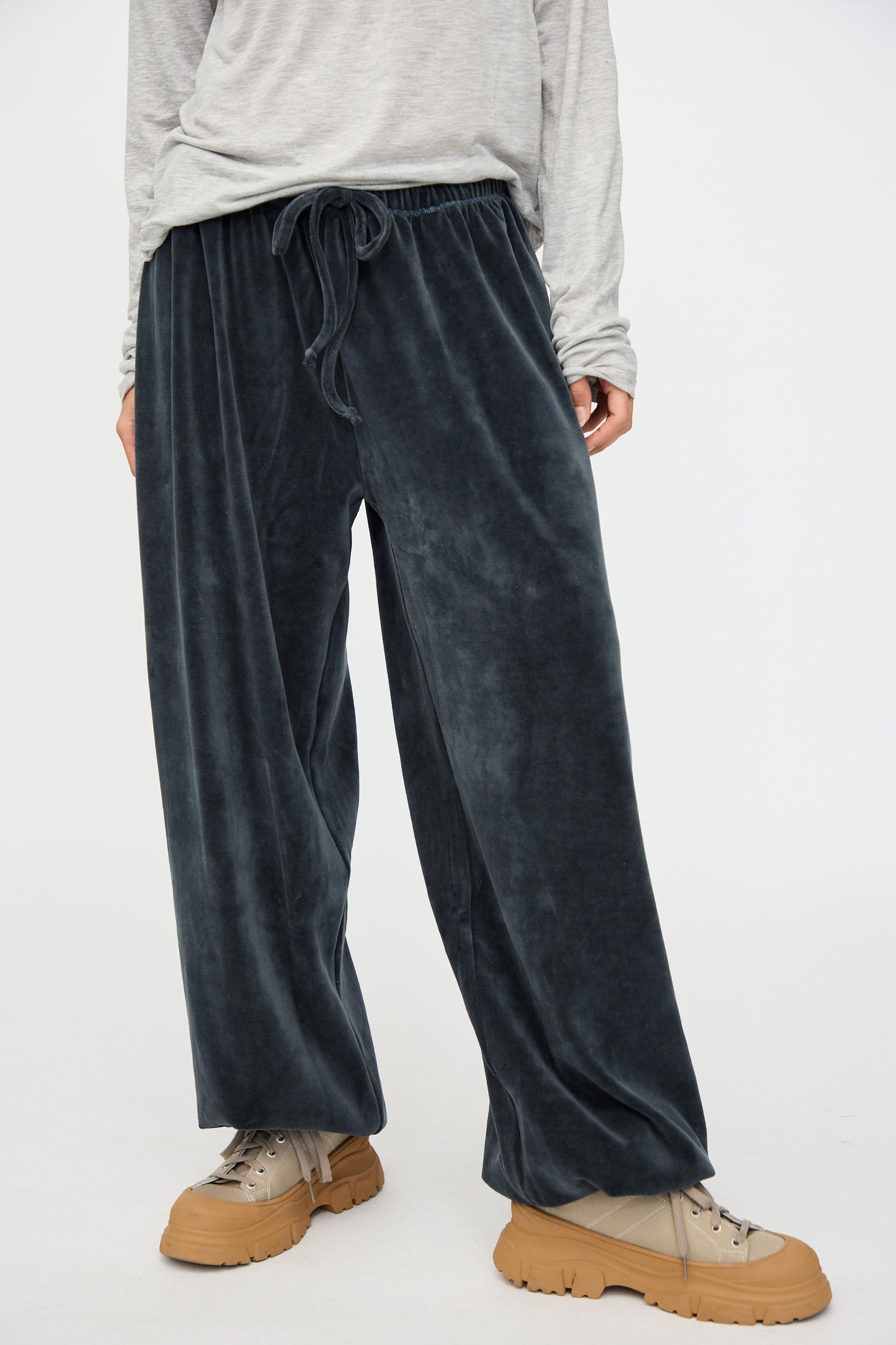 A person wears Baserange's Velour Pillow Pant in Petrolium Blue with a light gray long-sleeve top and tan chunky-soled shoes, standing against a plain light background.