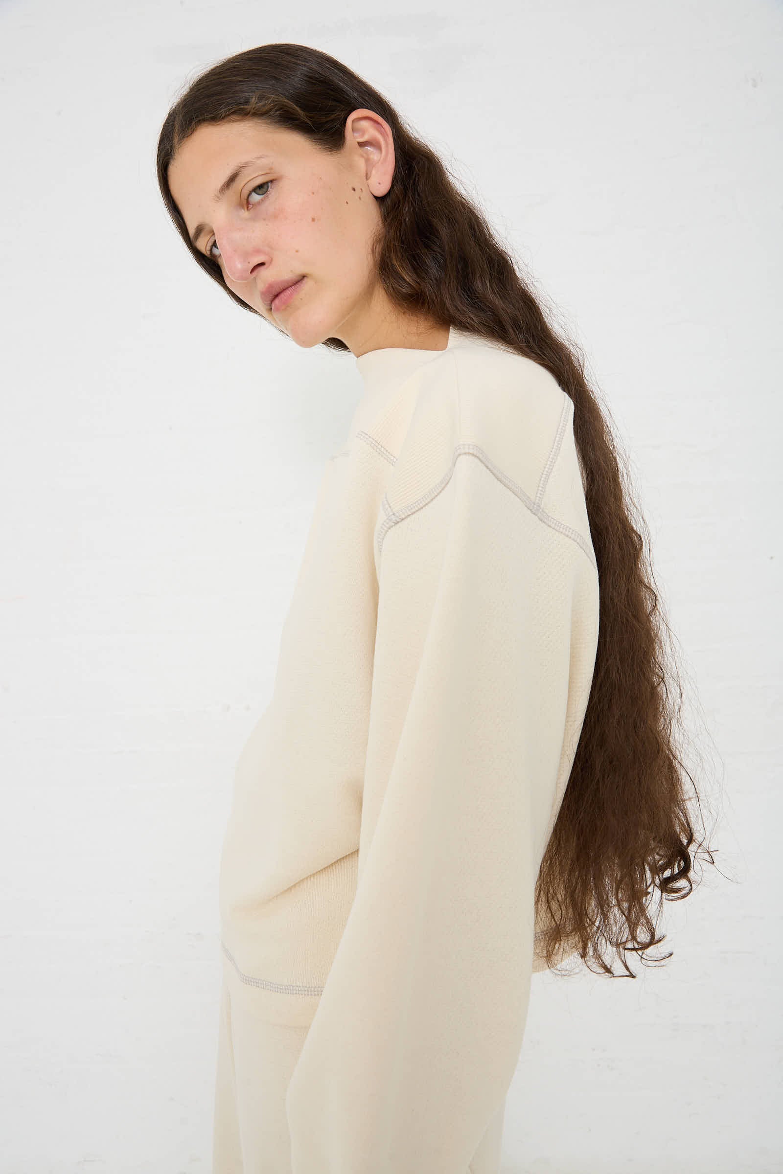 Person with long, dark hair stands against a plain white background, wearing a Way Boat Sweatshirt in Undyed Path Grey by Baserange made from regenerative cotton.