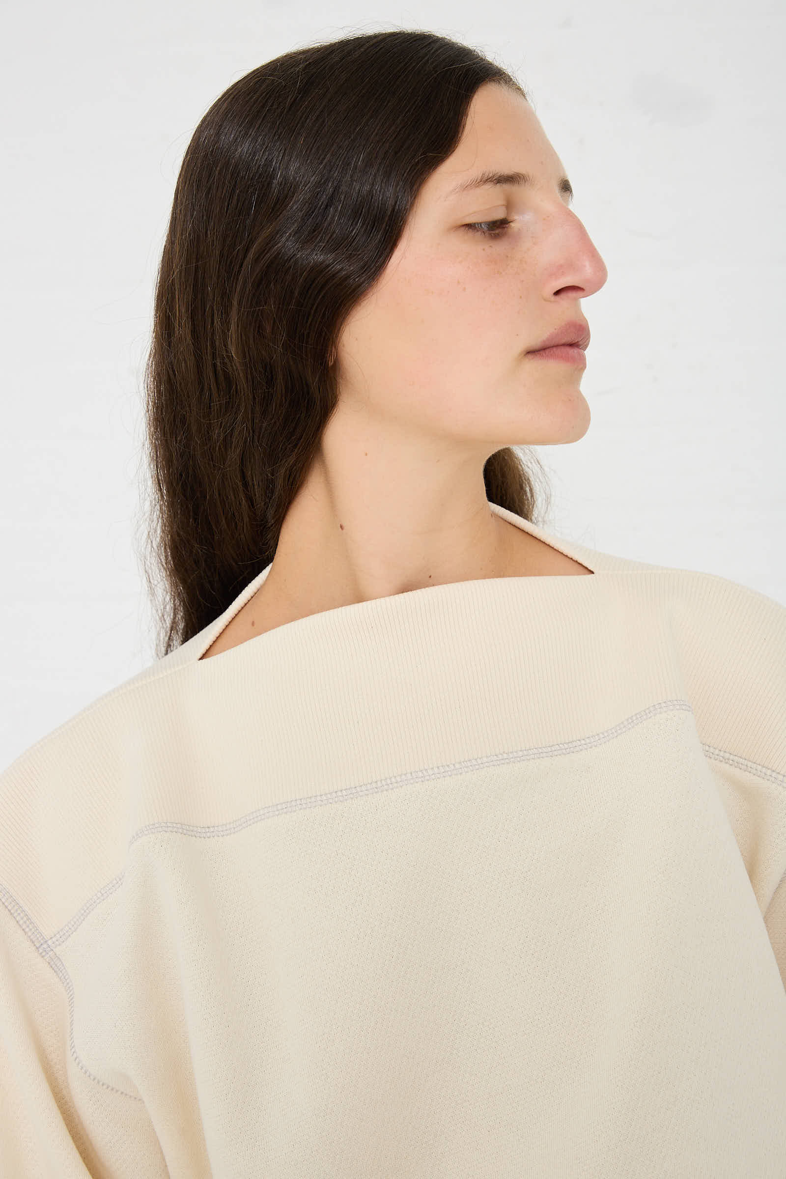 Person with long, dark hair, wearing a Way Boat Sweatshirt in Undyed Path Grey by Baserange, looking to the side against a plain background.