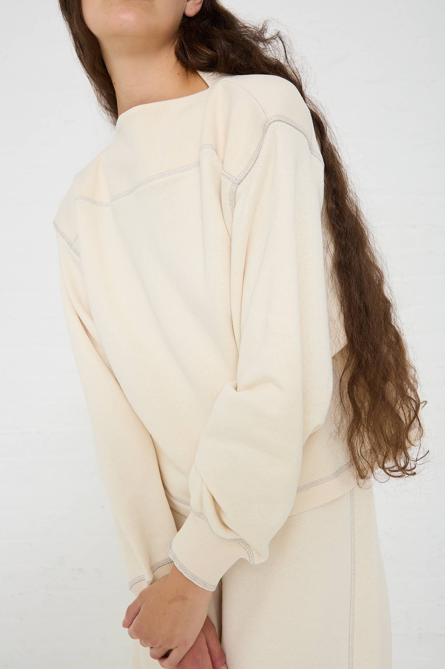 Person wearing a Baserange Way Boat Sweatshirt in Undyed Path Grey with long sleeves and matching pants, standing against a white background. Their long, wavy hair cascades over their shoulder.