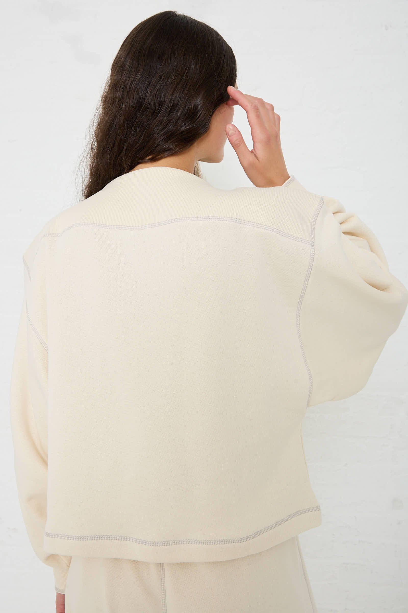 A person with long hair wearing a Baserange Way Boat Sweatshirt in Undyed Path Grey made from regenerative cotton faces away, with their left hand touching their head against a white background.