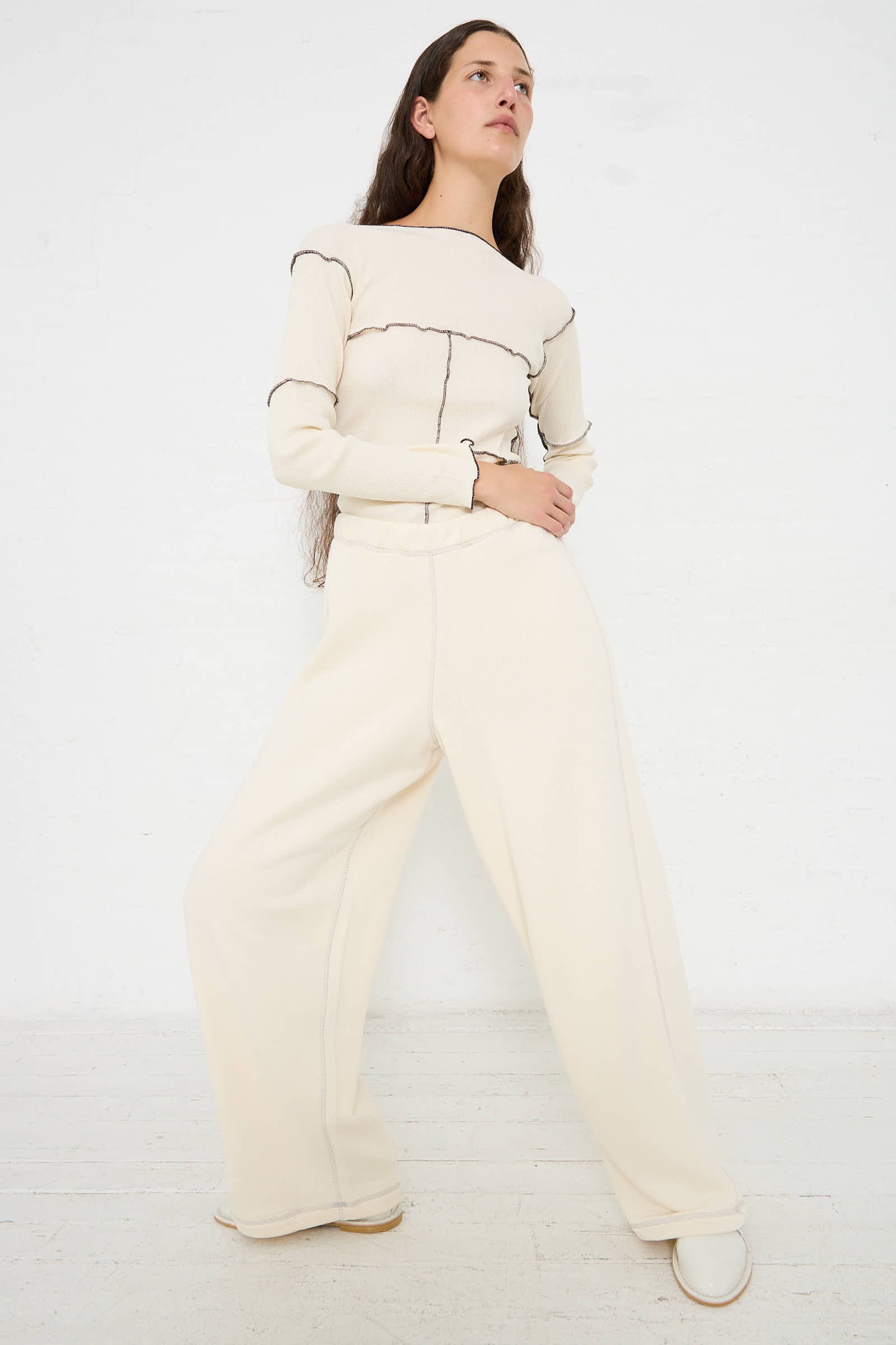 A person with long hair stands against a white background wearing the Baserange Way Pant in Undyed Path Grey, featuring black stitching and an elastic waistband for a relaxed fit, paired with white shoes.