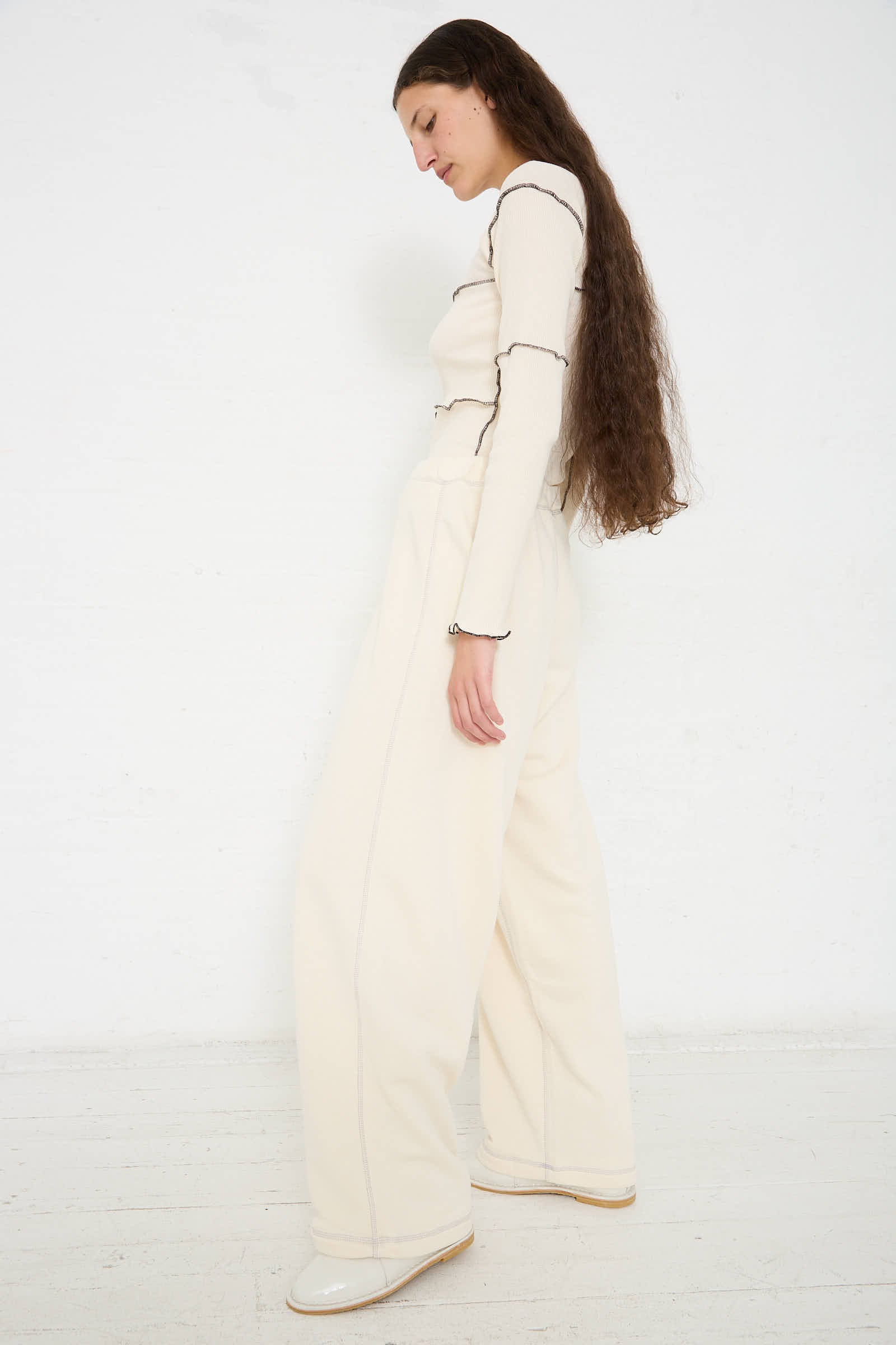 A person with long hair is standing sideways, wearing a **Baserange Way Pant in Undyed Path Grey** and white shoes, against a plain white background.