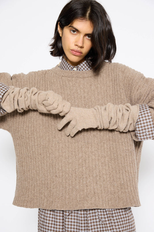 Person with short, dark hair wearing a loose beige knit sweater over a checkered shirt and matching Whit Gloves in Stoppa Brown by Baserange.