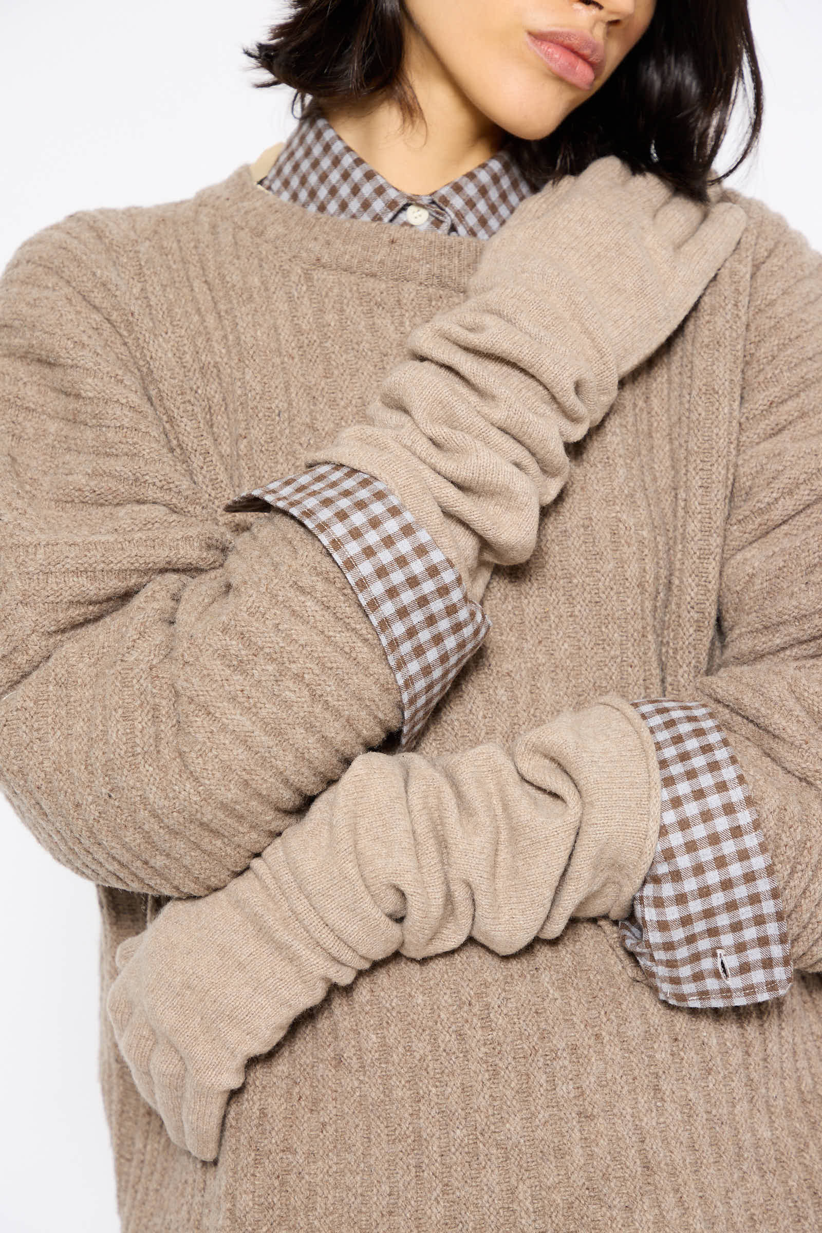 Close-up of a person wearing a textured brown sweater over a checkered shirt, with Baserange's Whit Gloves in Stoppa Brown, beige scrunched long knit gloves, covering their forearms. The head is partially visible, with features obscured.