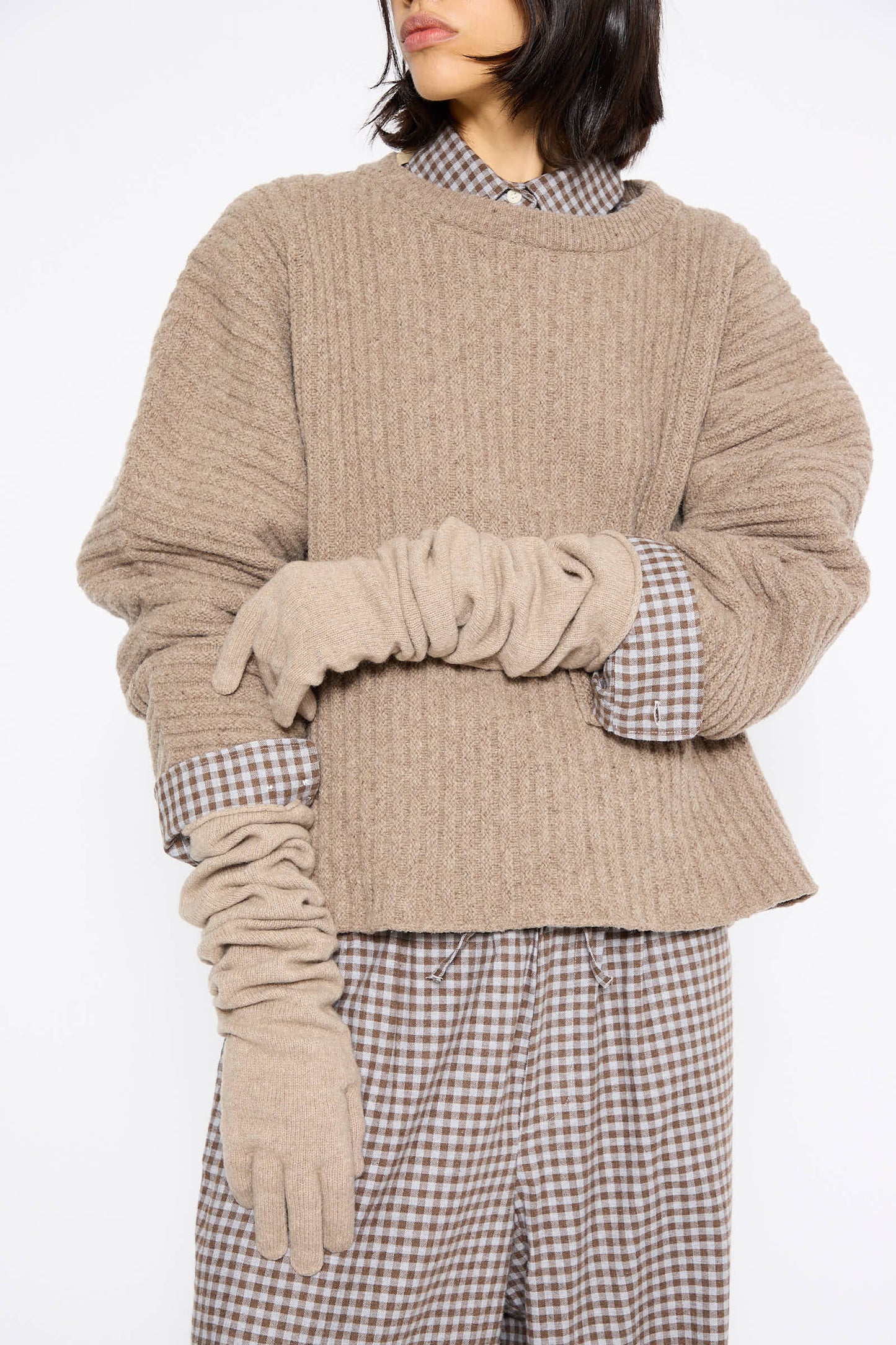 A person wearing a beige textured sweater made from recycled cashmere over a checkered shirt and pants, accessorized with Baserange's Whit Gloves in Stoppa Brown. The background is plain white.