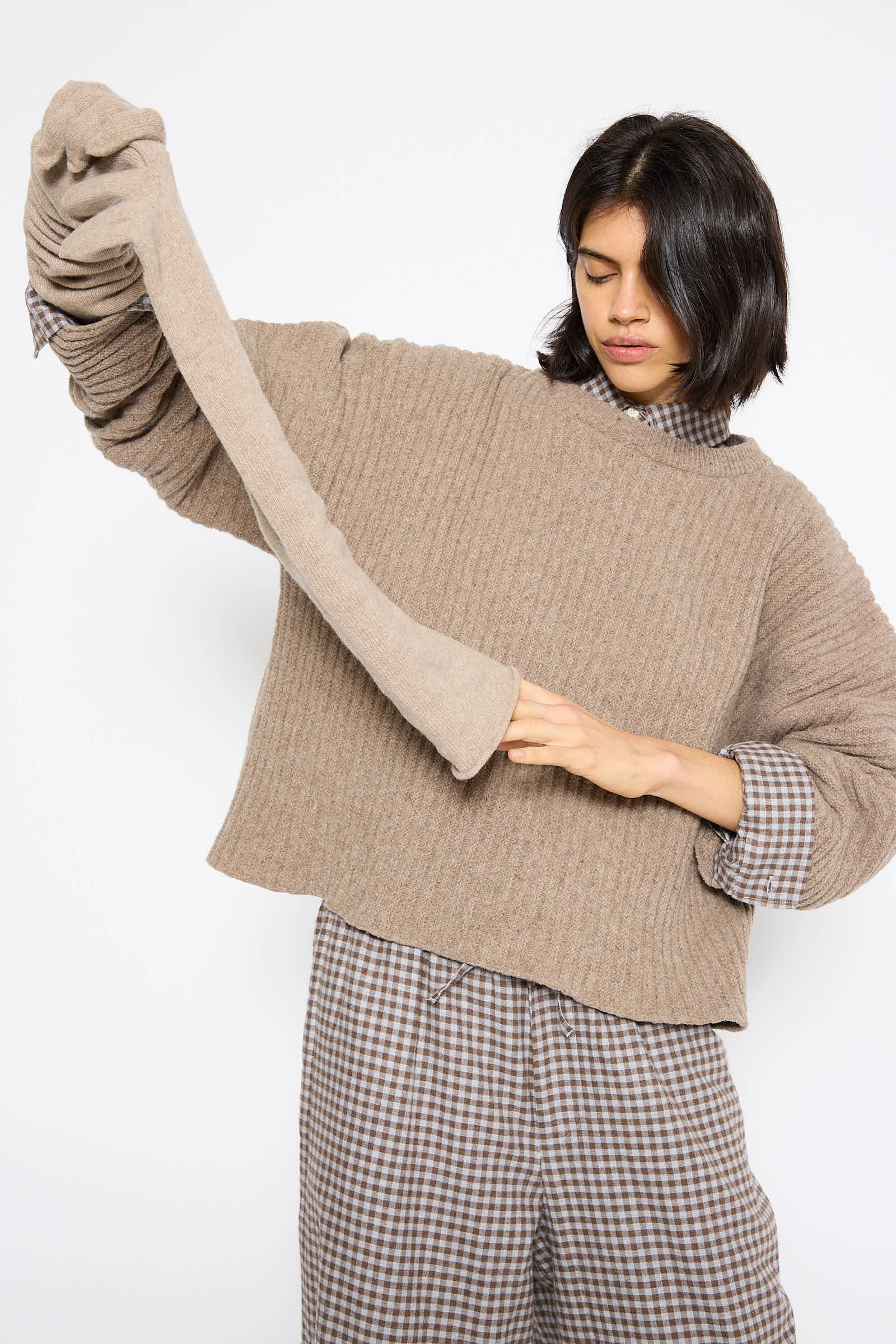 A person is wearing a beige sweater crafted from recycled cashmere and checkered pants, with an uncommonly long sleeve extending down to their knee. They seem to be adjusting this extended sleeve while sporting Whit Gloves in Stoppa Brown from Baserange.