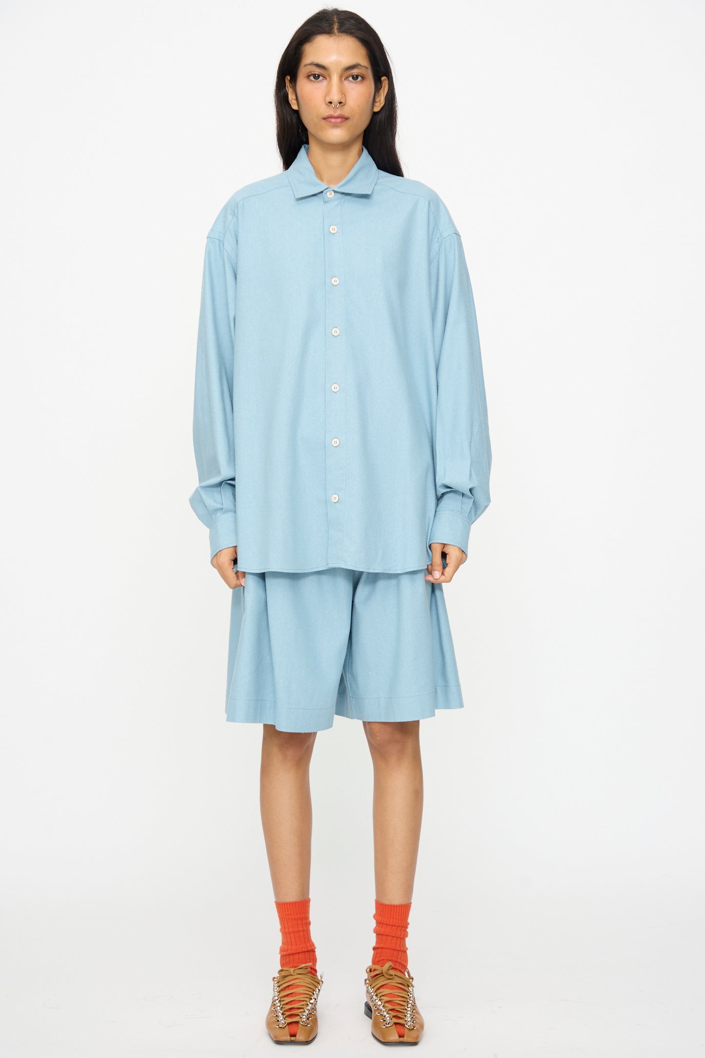A person faces forward in Baserange’s Wild Shorts in Seri Blue, styled with a light blue oversized button-up shirt, orange socks, and tan sandals, set against a plain white background.