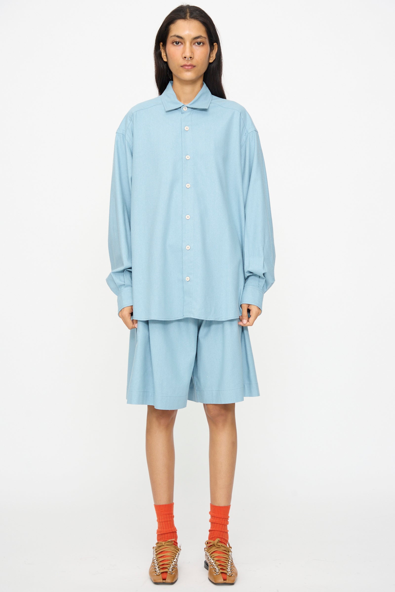 A person faces forward in Baserange’s Wild Shorts in Seri Blue, styled with a light blue oversized button-up shirt, orange socks, and tan sandals, set against a plain white background.
