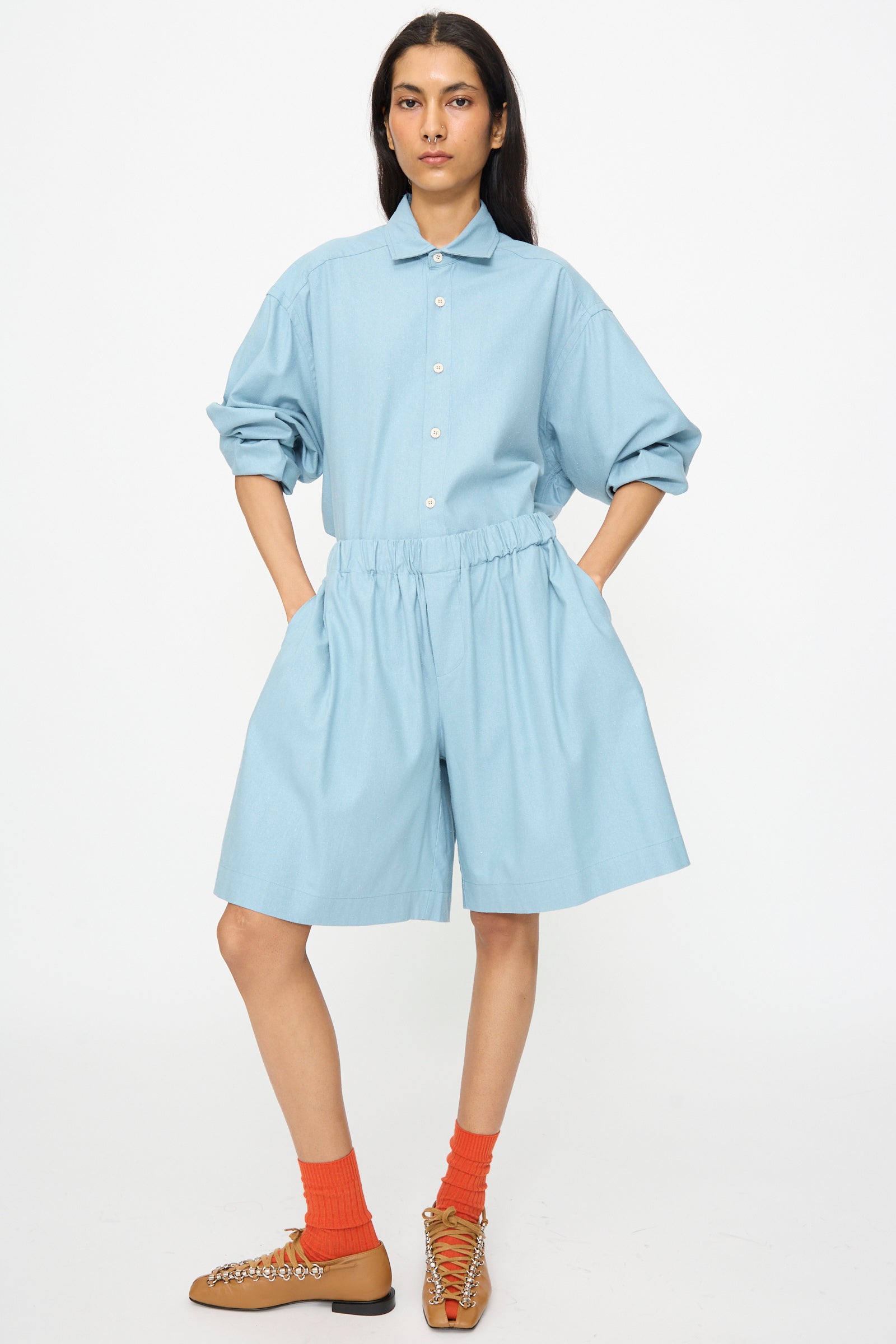 A person stands with hands in pockets, wearing Baserange Wild Shorts in Seri Blue, a light blue button-up shirt, orange socks, and tan shoes against a plain white background.