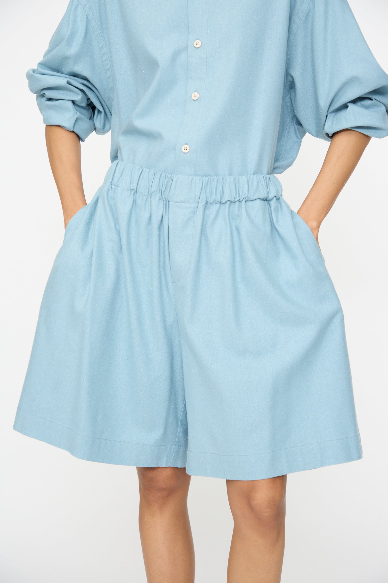 A person wears Baserange’s Wild Shorts in Seri Blue, a relaxed fit with an elasticated waistband, shown from shoulders to knees against a white background.