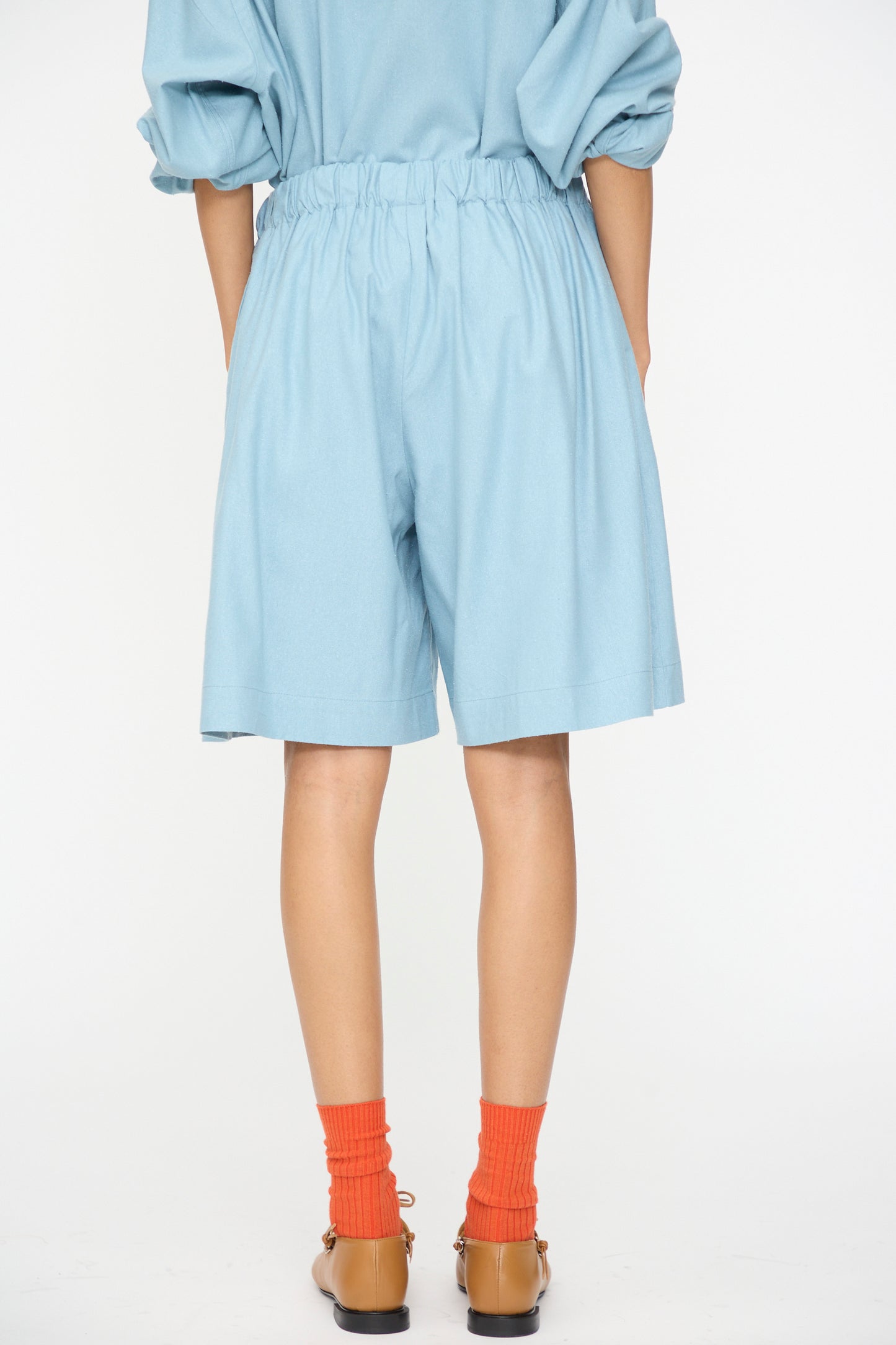 Model wears Baserange Wild Shorts in Seri Blue with an elastic waistband, paired with a matching untucked top, orange socks, and tan flats. Shown from the back against a plain white background.
