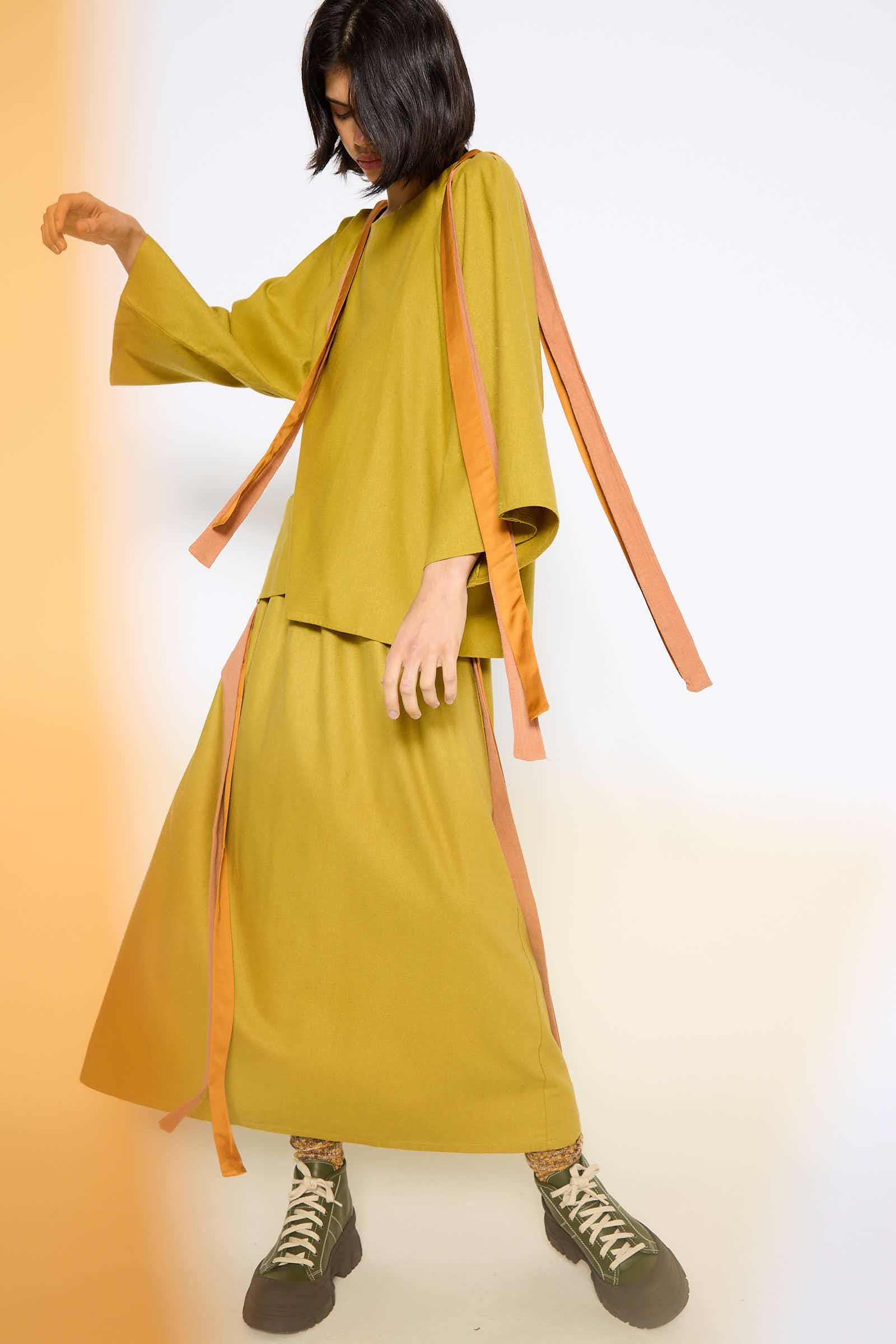 A person wearing an olive-green outfit with wide, long sleeves and orange ribbons poses against a plain white background. They have short hair, wear dark sneakers, and sport the Wild Silk Cena Skirt in Sophos Green by Baserange, featuring an elasticated waist.