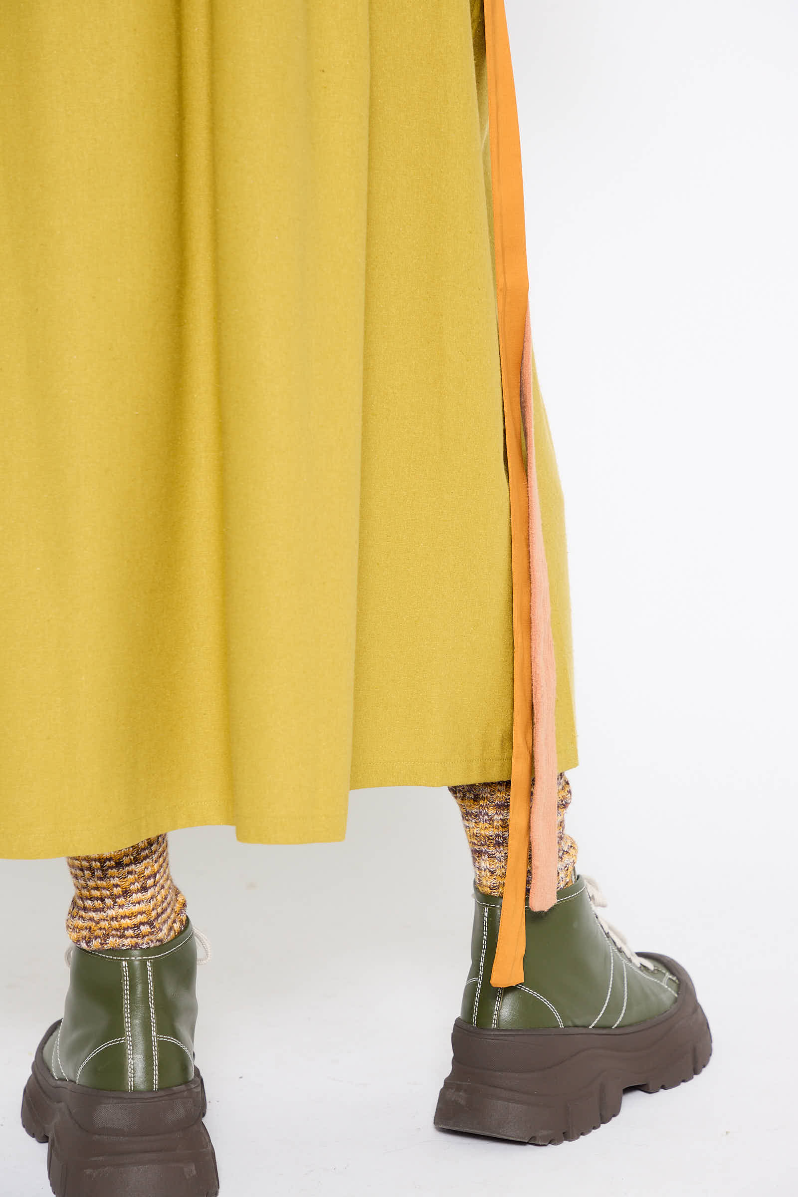 Close-up of a person wearing green platform shoes, patterned socks, and the Baserange Wild Silk Cena Skirt in Sophos Green with an orange ribbon detail and elasticated waist. The background is plain white.