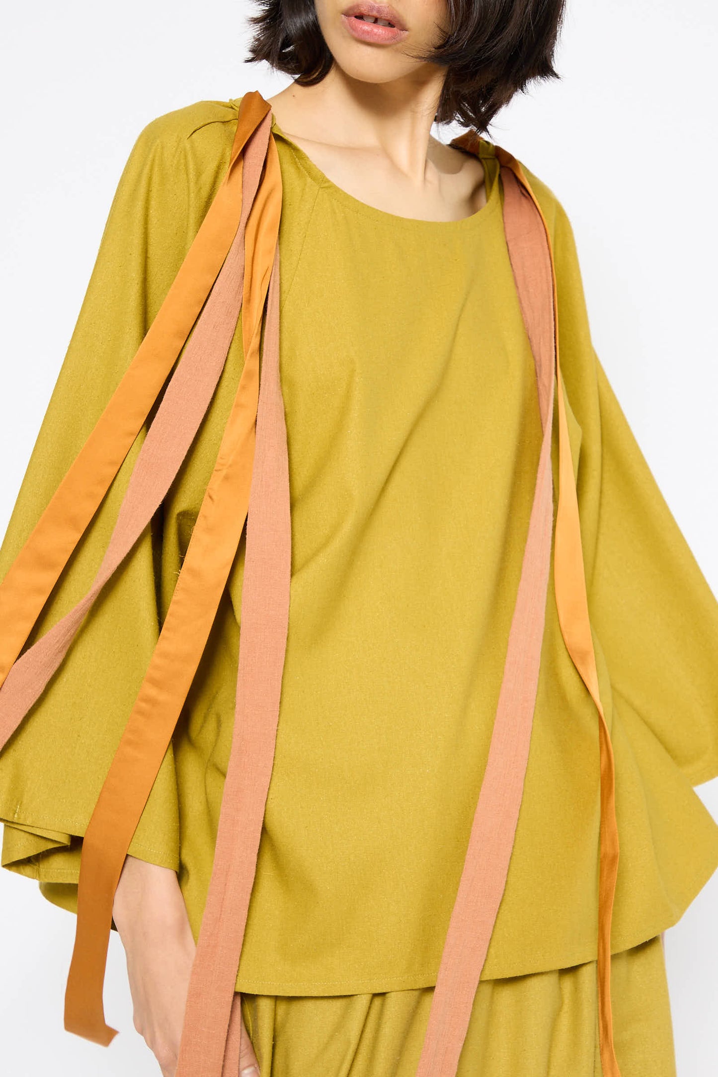 Person wearing a loose-fitting, Sophos Green Wild Silk Cena Top by Baserange, adorned with long, orange and brown ribbons. The blouse features 3/4 sleeves, and only the upper body is visible against a plain white background.