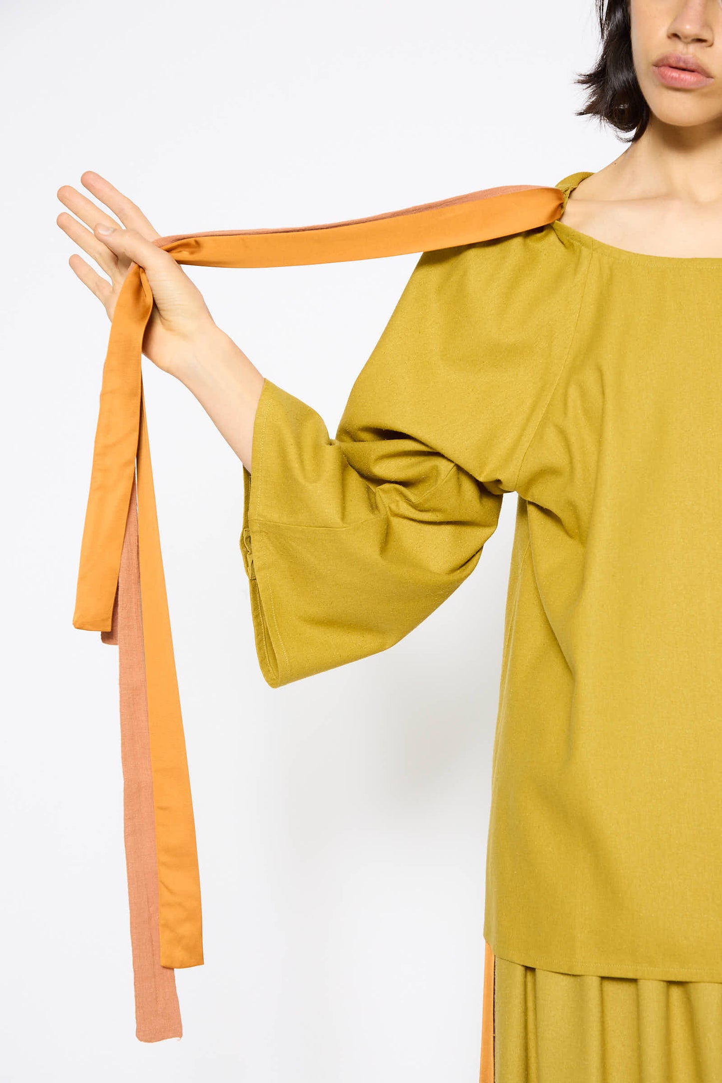 A person in a Wild Silk Cena Top in Sophos Green by Baserange holds orange fabric ties attached to their shoulder. Their face is partially visible, and the background is plain white.