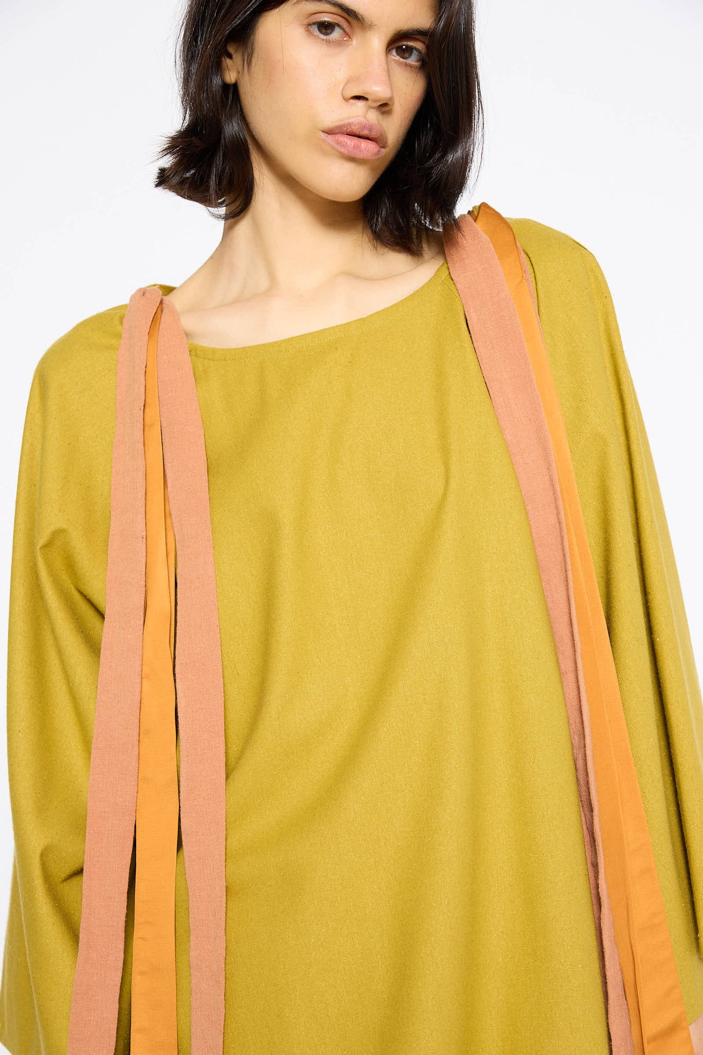 A person with shoulder-length dark hair wearing a Sophos Green Wild Silk Cena Top by Baserange, featuring orange and pink fabric strips draped over their shoulders.