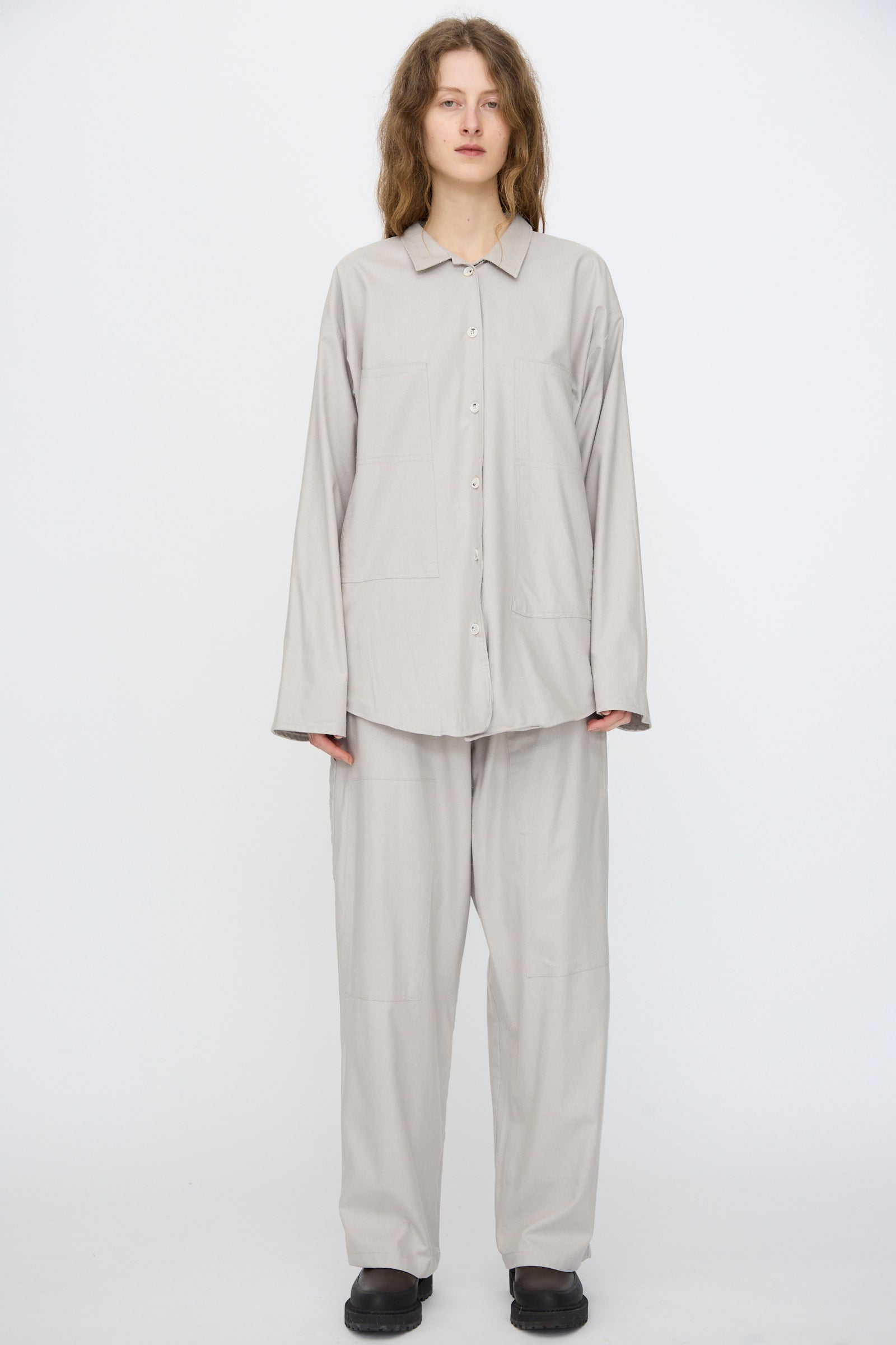A person stands against a plain white background wearing the Baserange Wild Silk Gren Overshirt in Prun Grey, styled with matching pants and black shoes.