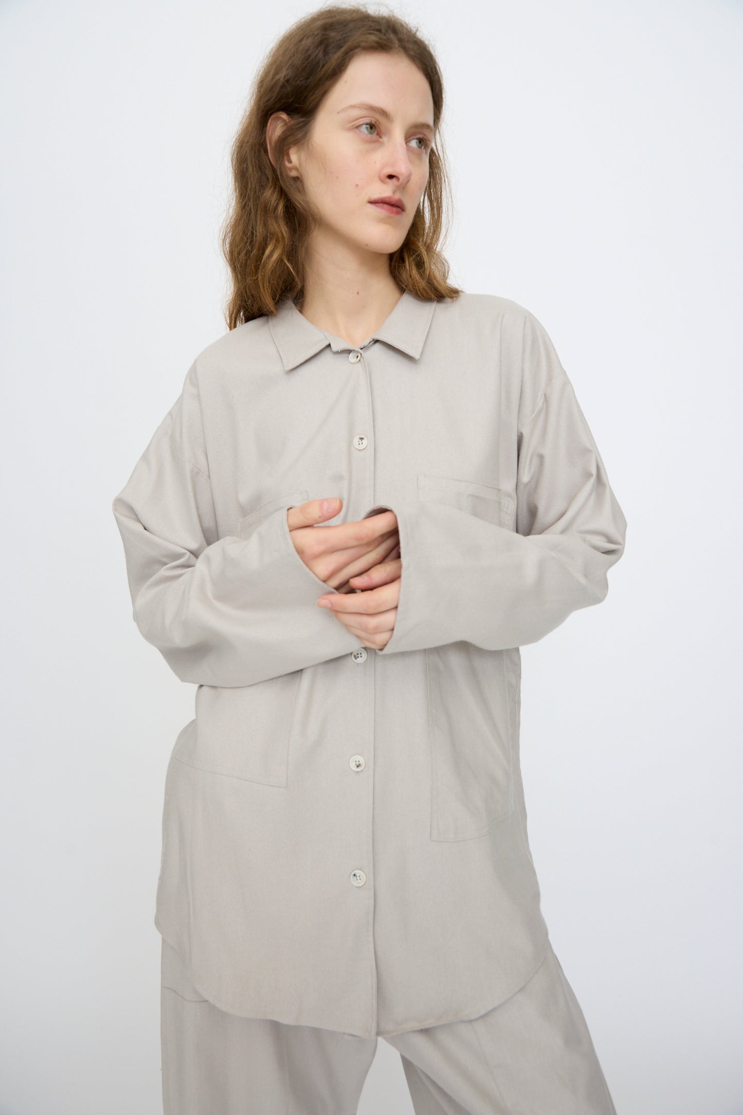 A person with wavy hair wears the Baserange Wild Silk Gren Overshirt in Prun Grey—oversized, made in Türkiye—paired with matching pants, standing against a plain white background.
