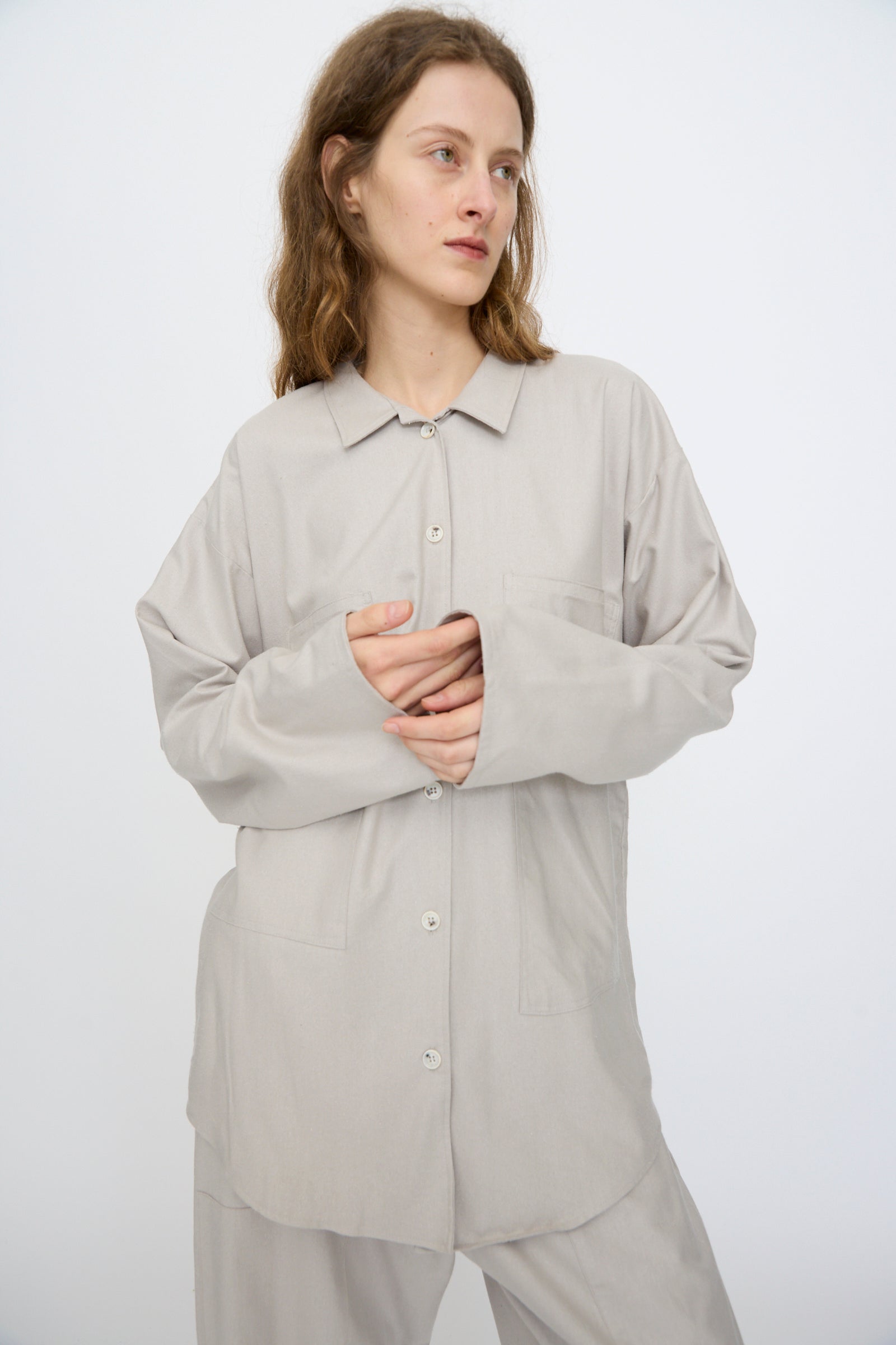 A person with wavy hair wears the Baserange Wild Silk Gren Overshirt in Prun Grey—oversized, made in Türkiye—paired with matching pants, standing against a plain white background.
