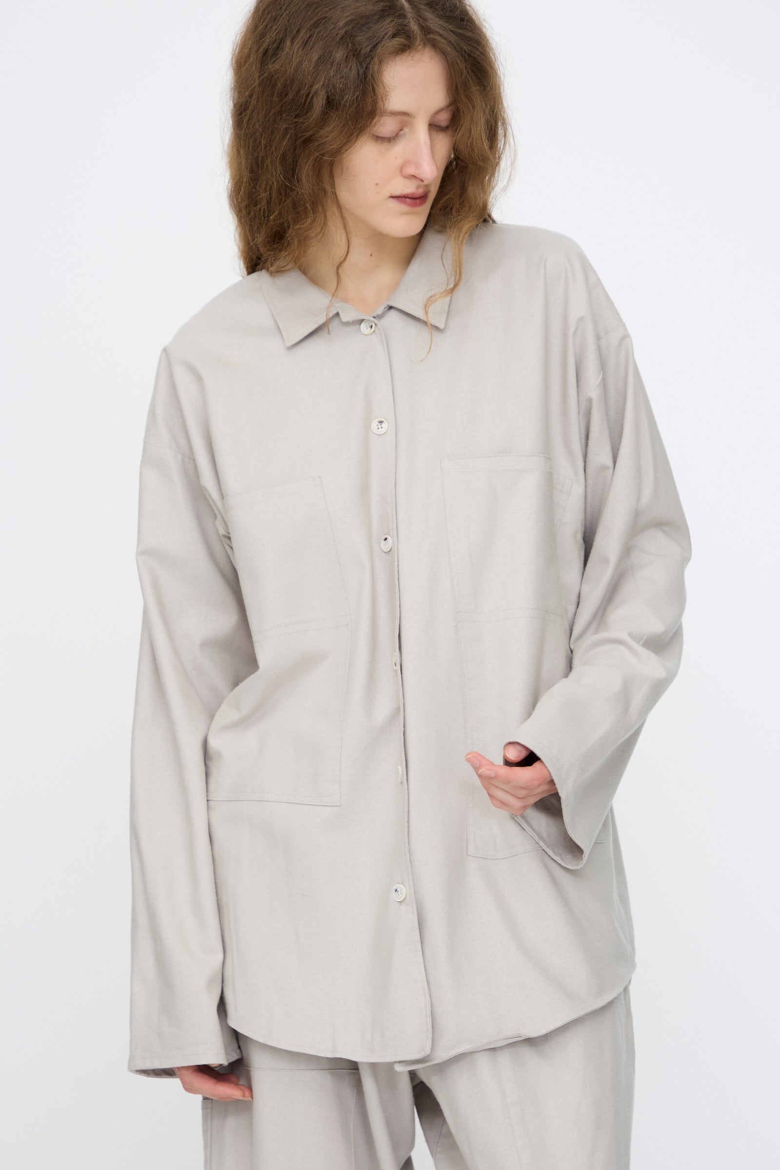 A person with wavy hair wears the Baserange Wild Silk Gren Overshirt in Prun Grey, made in Türkiye, and matching pants, standing against a plain white background.