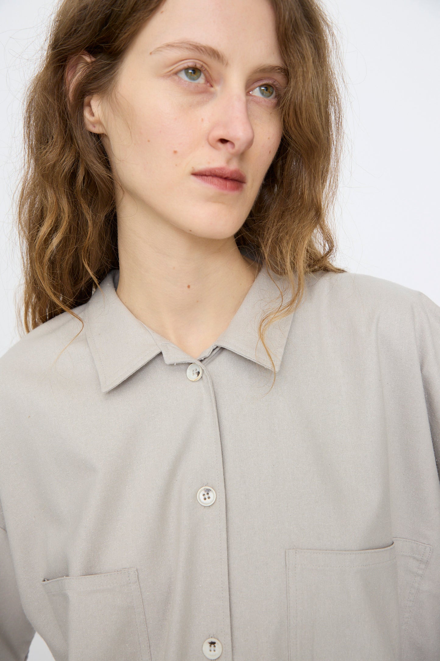 A person with long wavy hair wears the Baserange Wild Silk Gren Overshirt in Prun Grey, featuring two front pockets, and looks slightly to the side against a plain background.