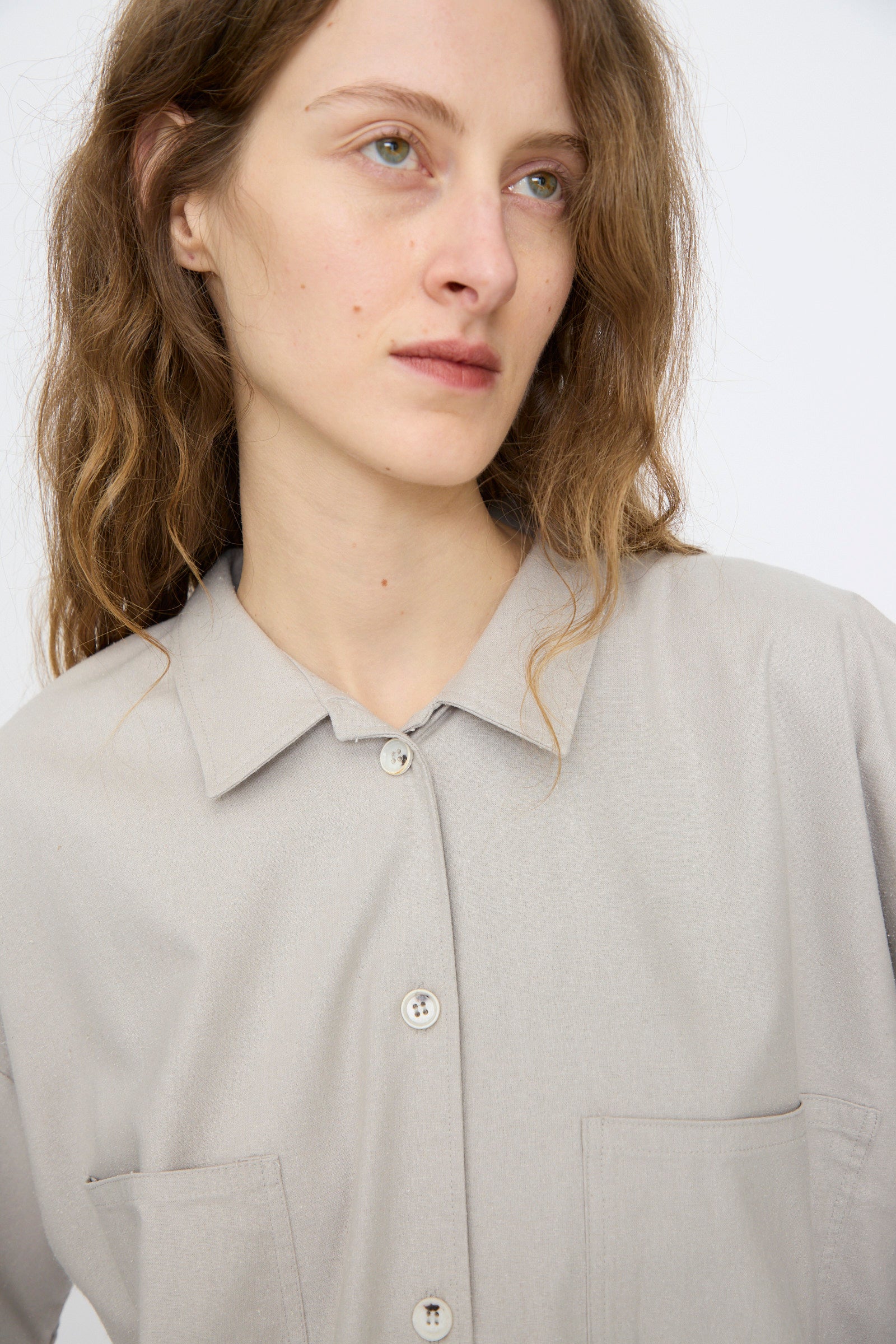 A person with long wavy hair wears the Baserange Wild Silk Gren Overshirt in Prun Grey, featuring two front pockets, and looks slightly to the side against a plain background.