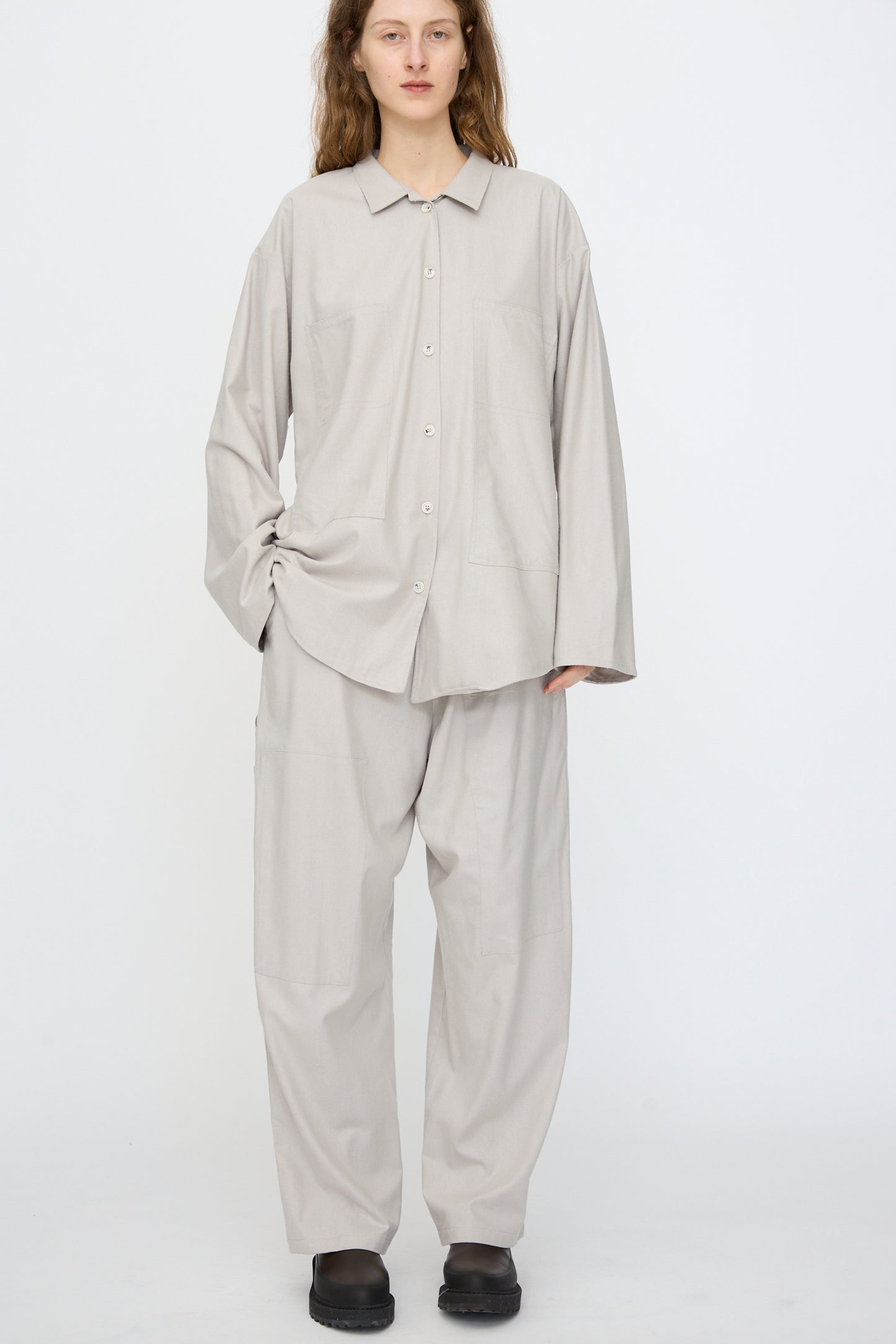 A person stands against a plain white background wearing a relaxed fit light gray button-up shirt, Baserange Wild Silk Gren Pant in Prun Grey with an elasticated waistband, and black shoes.