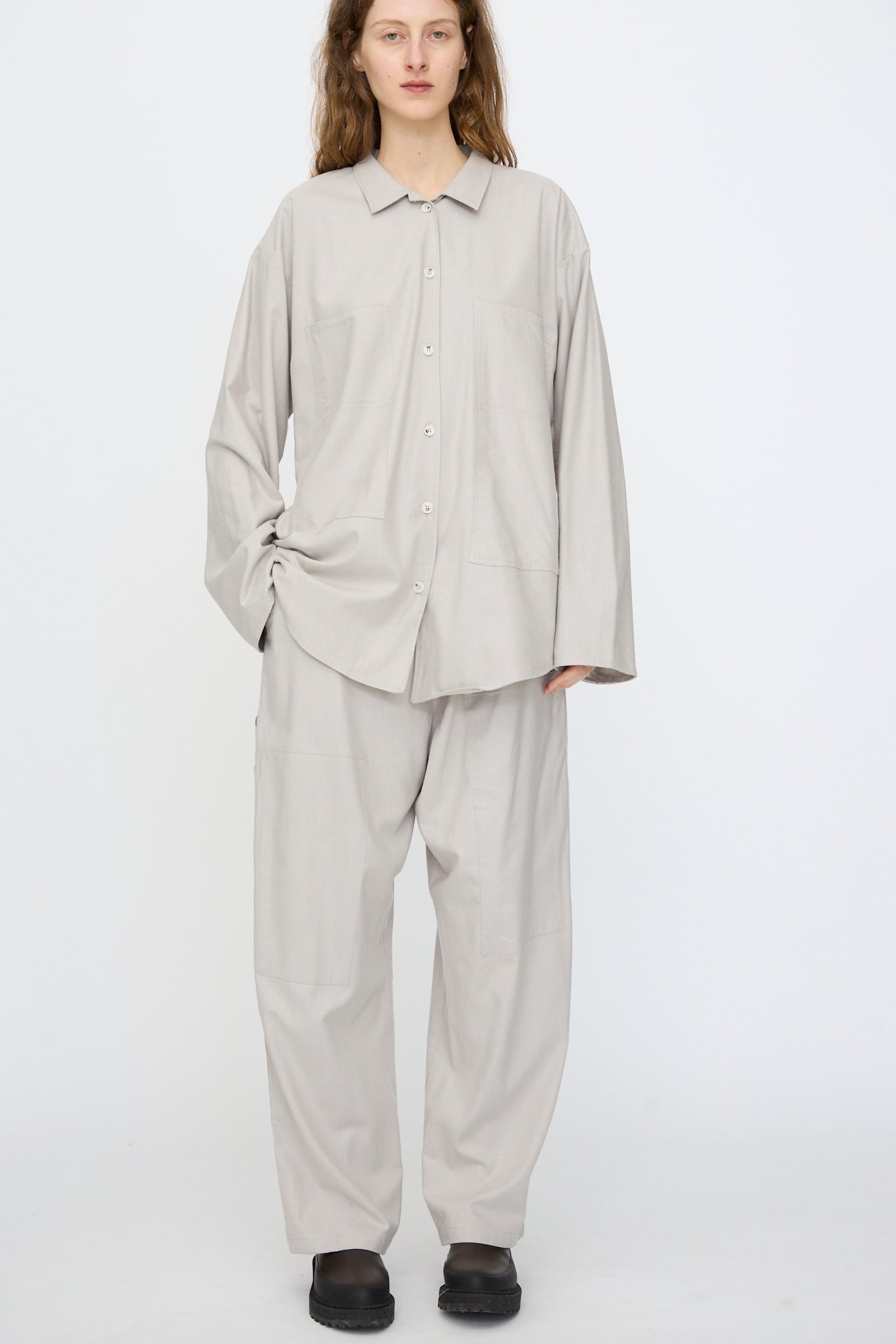 A person stands against a plain white background wearing a relaxed fit light gray button-up shirt, Baserange Wild Silk Gren Pant in Prun Grey with an elasticated waistband, and black shoes.