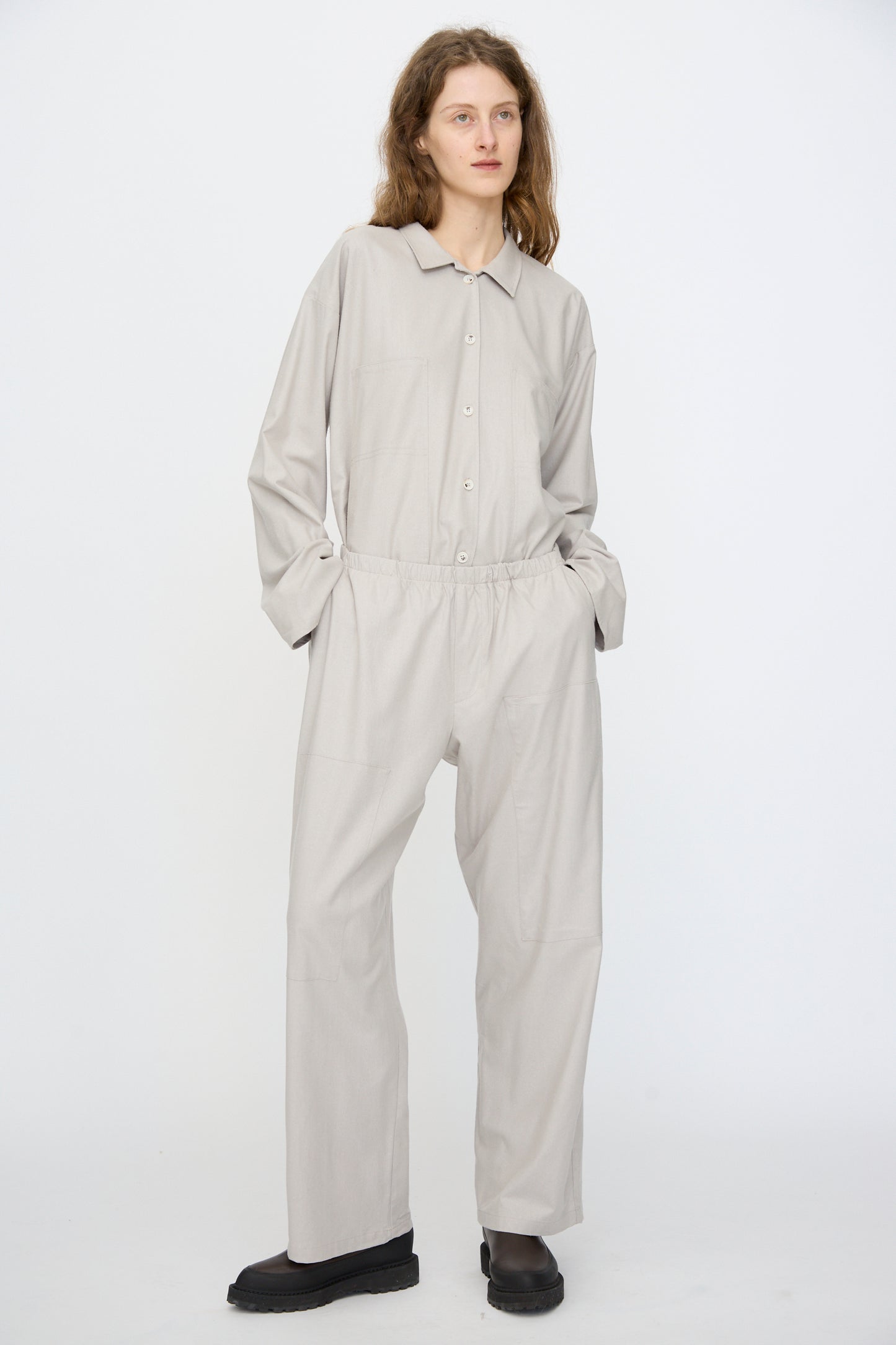 A person wears the Baserange Wild Silk Gren Pant in Prun Grey, styled with black shoes, standing against a white backdrop with hands in pockets and gazing slightly to the side.