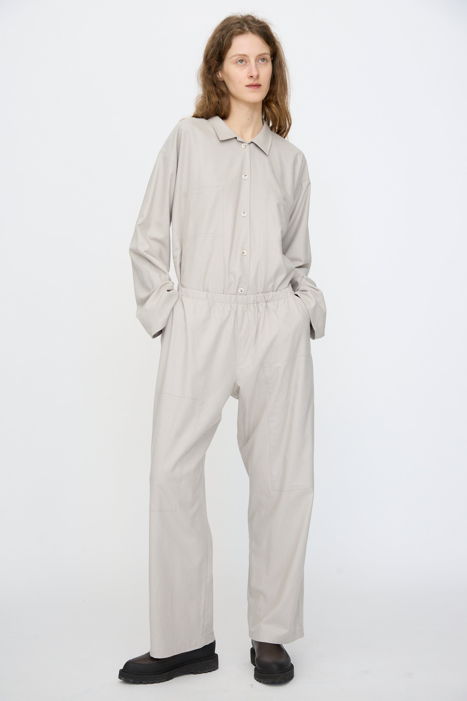 A person wears the Baserange Wild Silk Gren Pant in Prun Grey, styled with black shoes, standing against a white backdrop with hands in pockets and gazing slightly to the side.