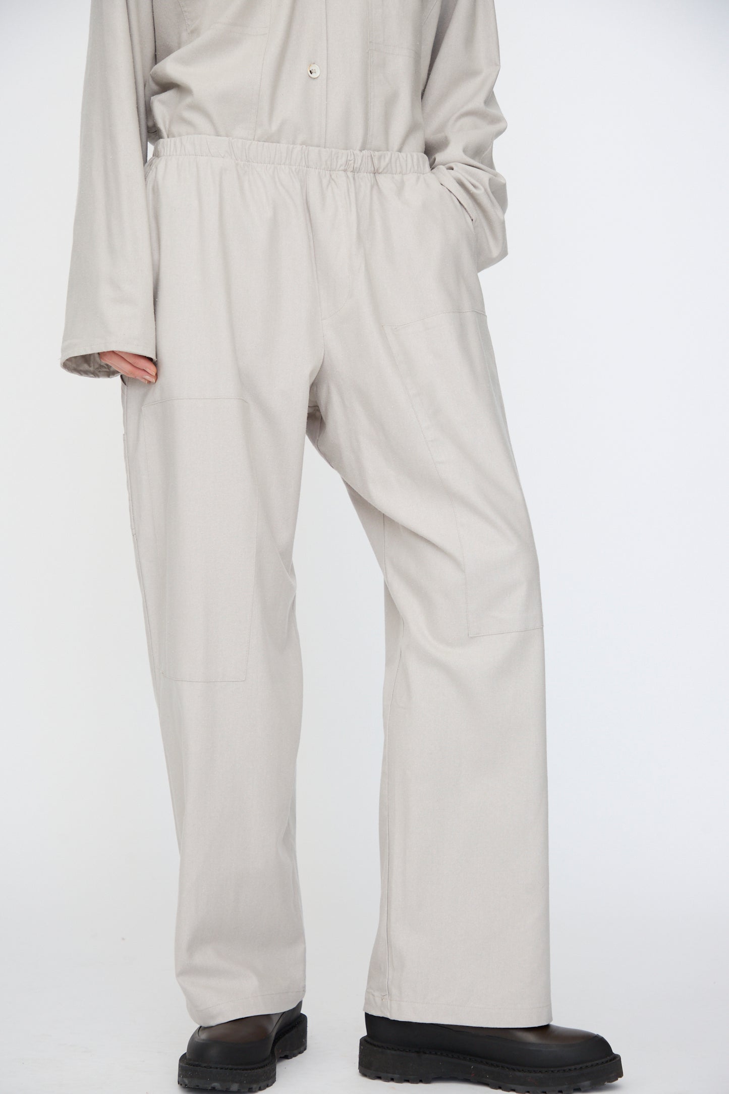 A person wearing Baserange’s Wild Silk Gren Pant in Prun Grey, featuring a relaxed fit and elasticated waistband, paired with a matching long-sleeve top and black shoes, shown from the shoulders down.