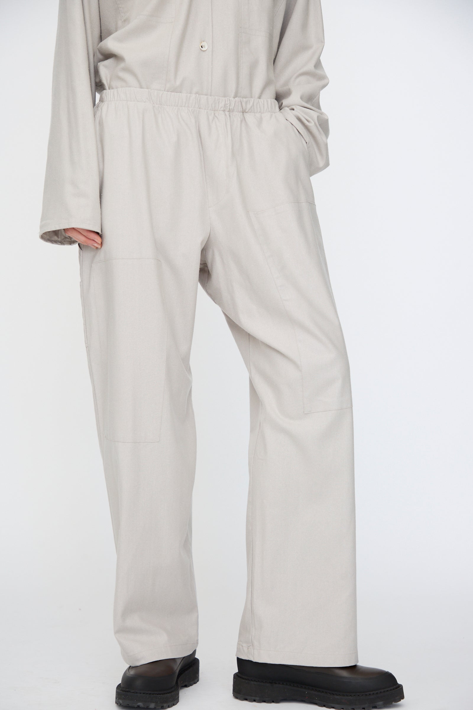 A person wearing Baserange’s Wild Silk Gren Pant in Prun Grey, featuring a relaxed fit and elasticated waistband, paired with a matching long-sleeve top and black shoes, shown from the shoulders down.