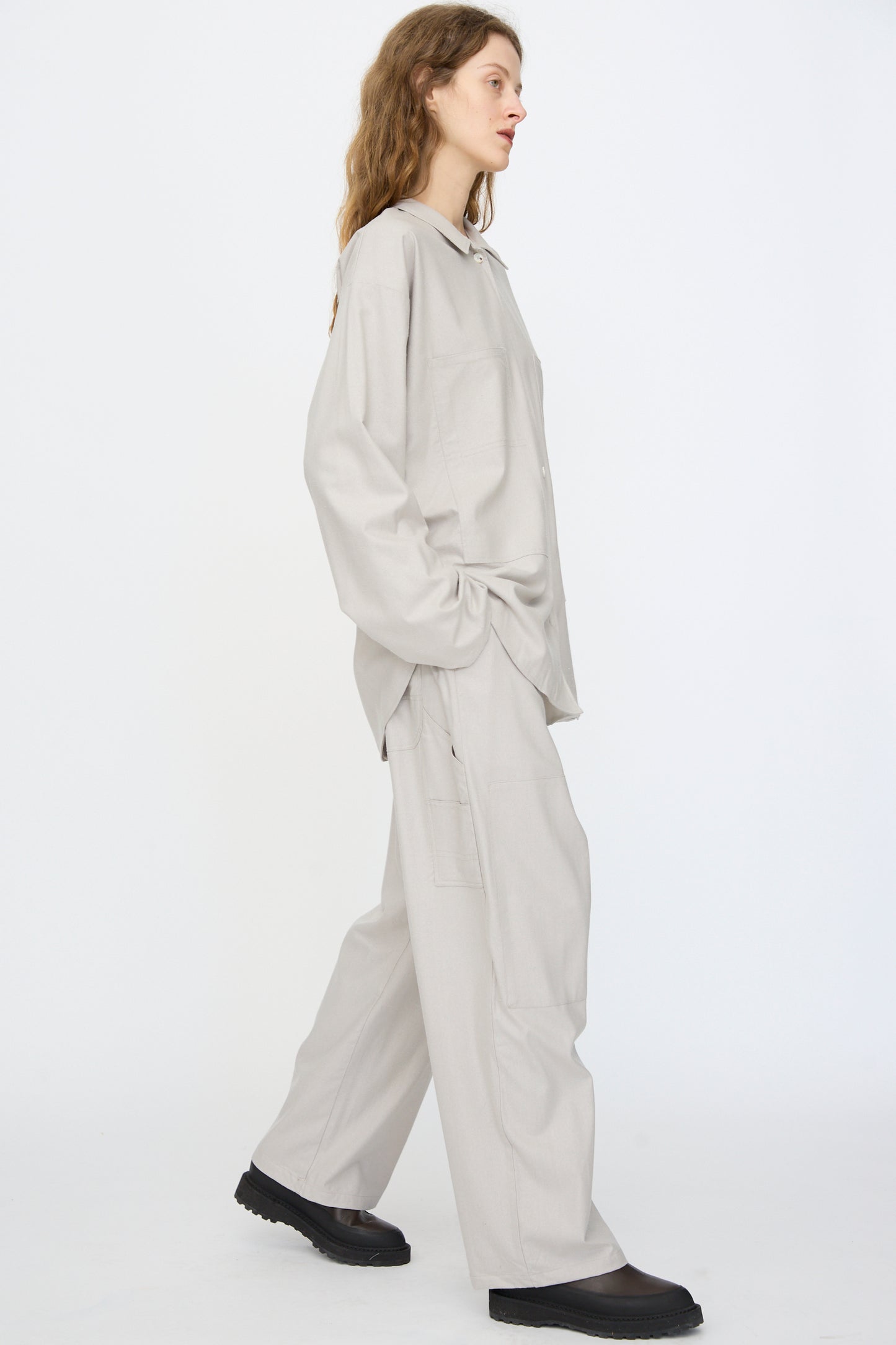 Person in profile wears an oversized light grey button-up shirt, Baserange Wild Silk Gren Pant in Prun Grey with elastic waistband, and black shoes, standing against a plain white background.