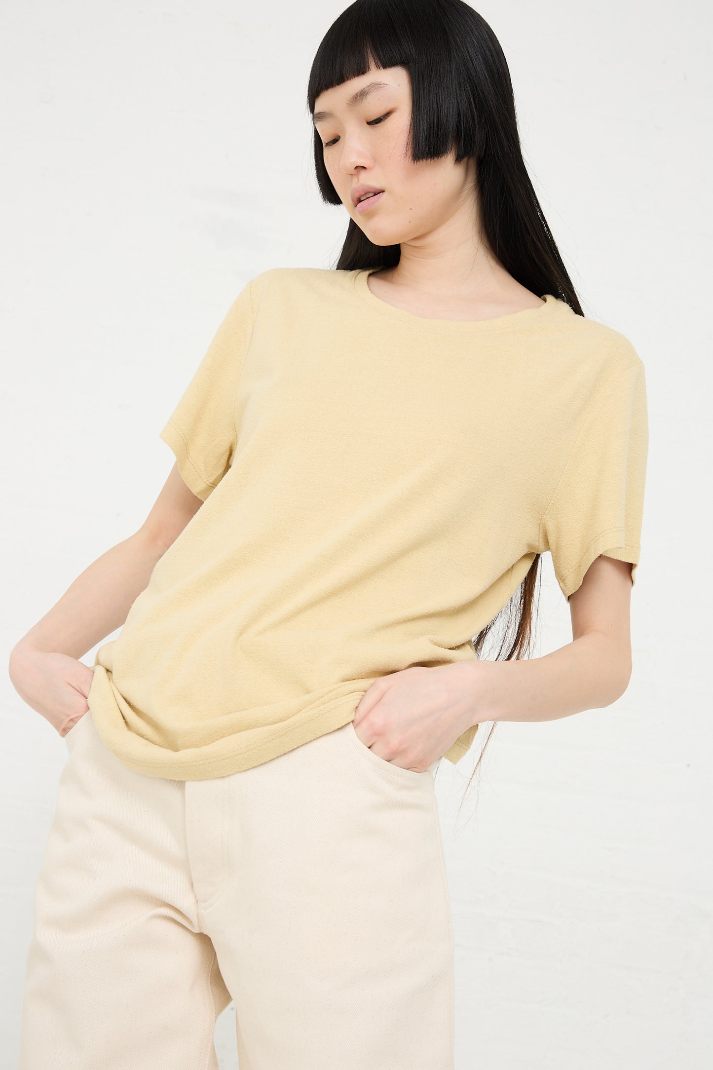 A woman with dark hair posing with her hand on her hip, wearing a Baserange Wild Silk Jersey Tee in Oak Yellow and beige pants against a white background.