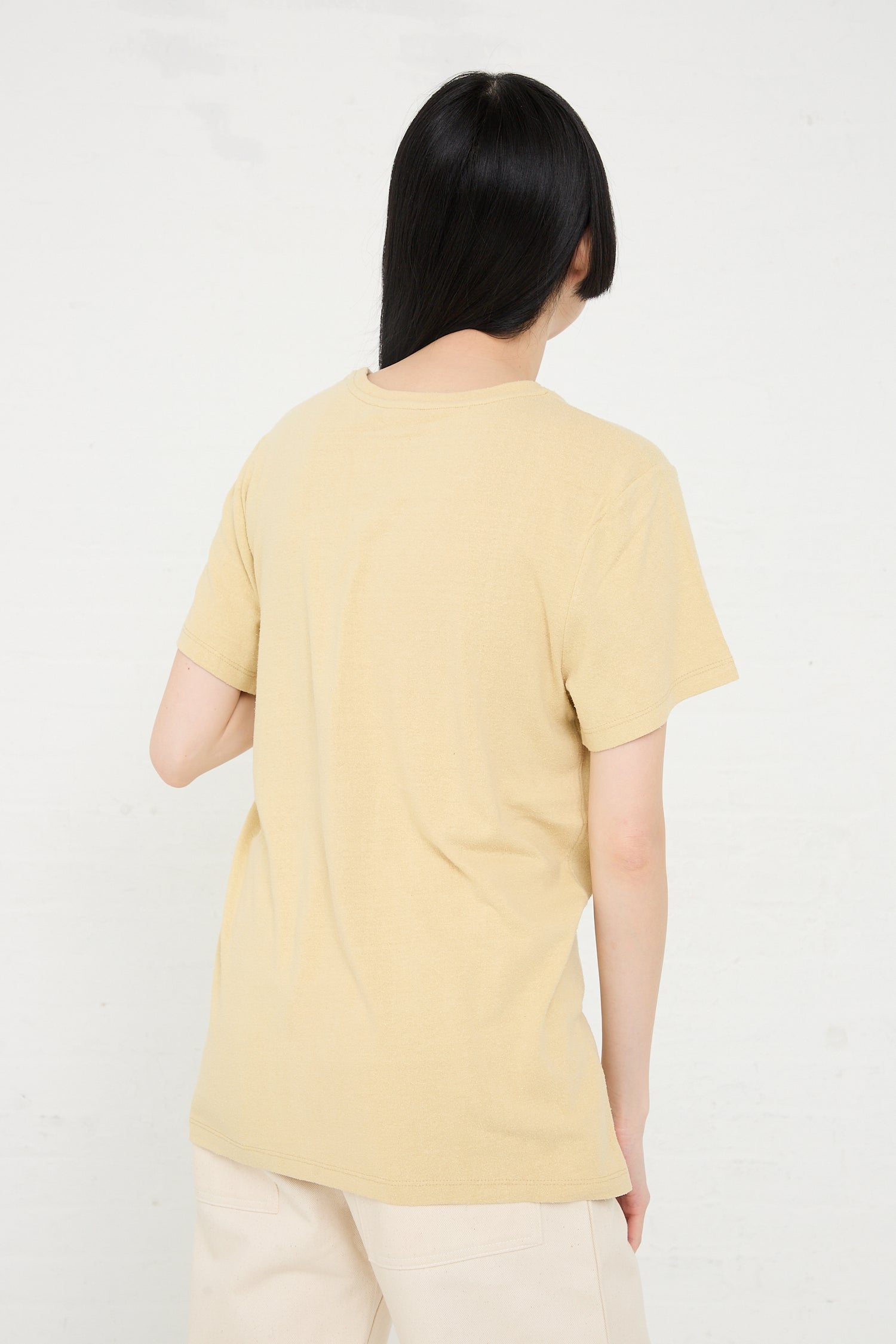 Woman from behind wearing a Baserange Wild Silk Jersey Tee in Oak Yellow sustainably produced and beige pants against a white background.