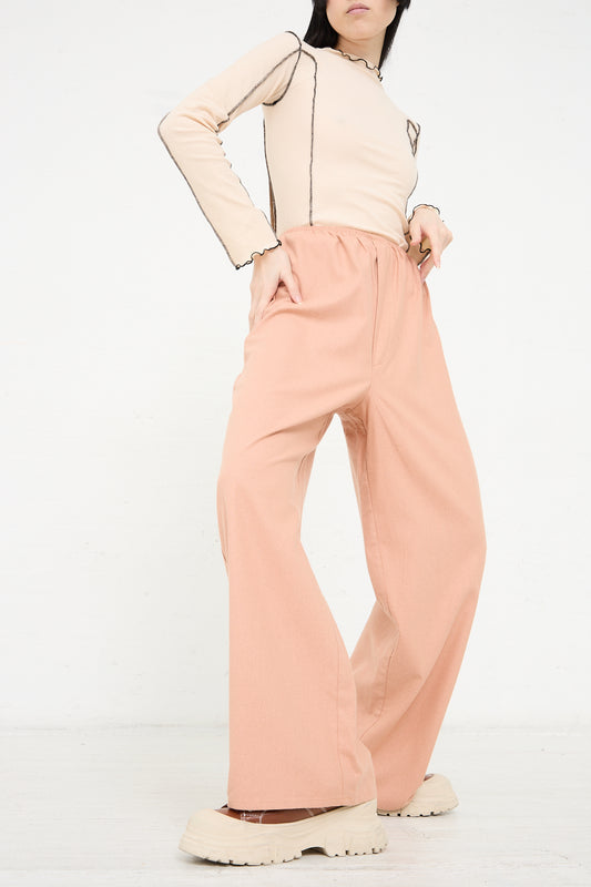 A person standing in a neutral pose wearing the Baserange Wild Silk Stoa Pant in Sid Pink, a beige long-sleeve top made of wild silk, and chunky beige shoes.