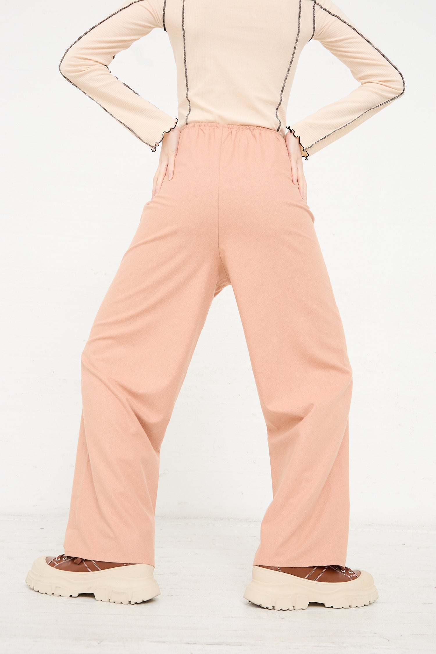 A person standing with hands on hips wearing the Wild Silk Stoa Pant in Sid Pink by Baserange and a beige top with chunky beige shoes, viewed from the back.
